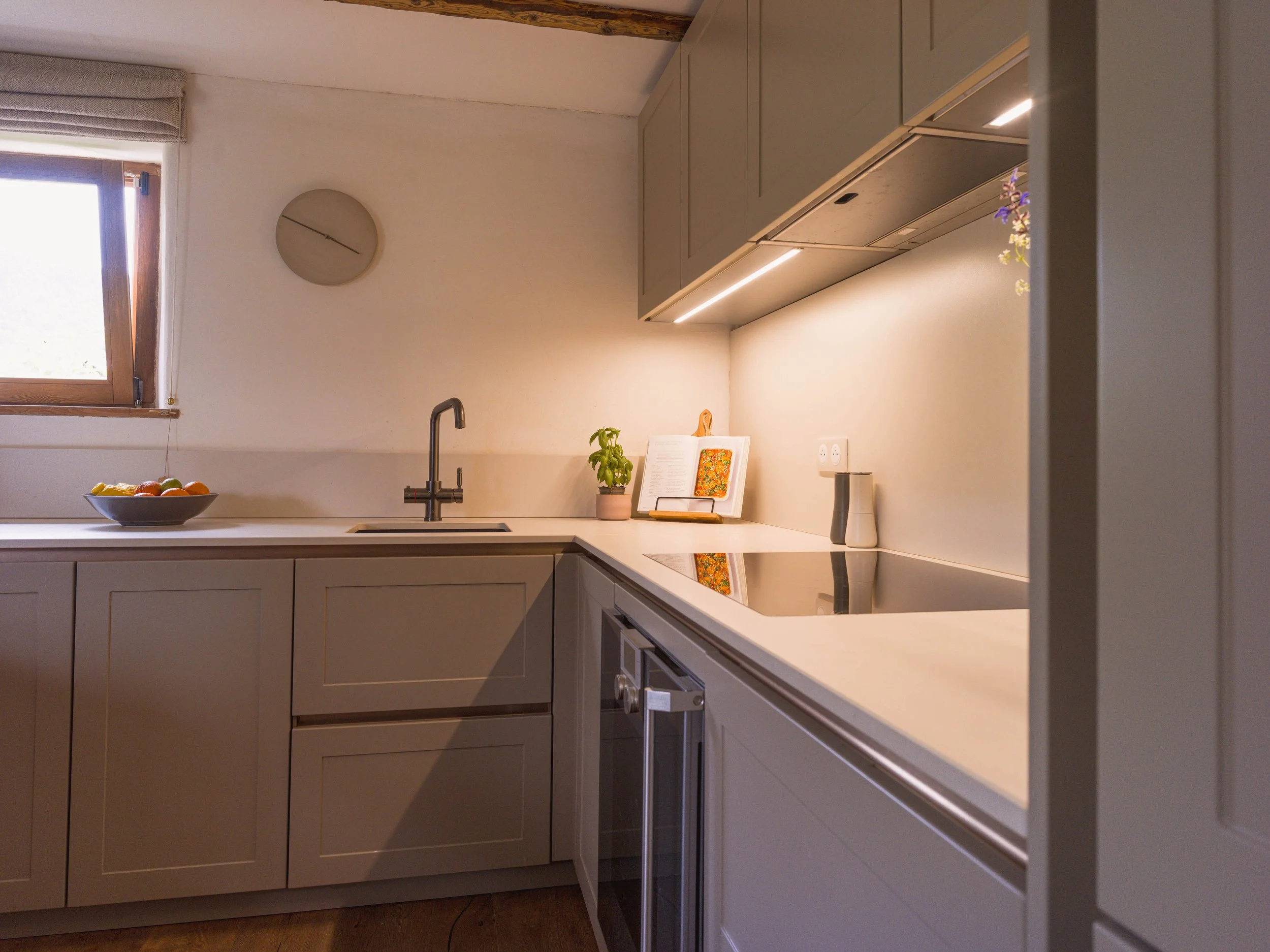 A modern kitchen with beige cabinets, a white countertop, and a window with wooden trim. On the counter, there is a bowl of various fruits, a small potted plant, and a cookbook stand with a book. The kitchen has under-cabinet lighting, a wall clock, 