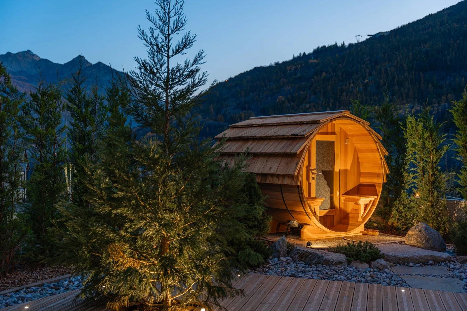 A wooden barrel-shaped outdoor sauna illuminated at dusk, surrounded by trees and mountains in the background.