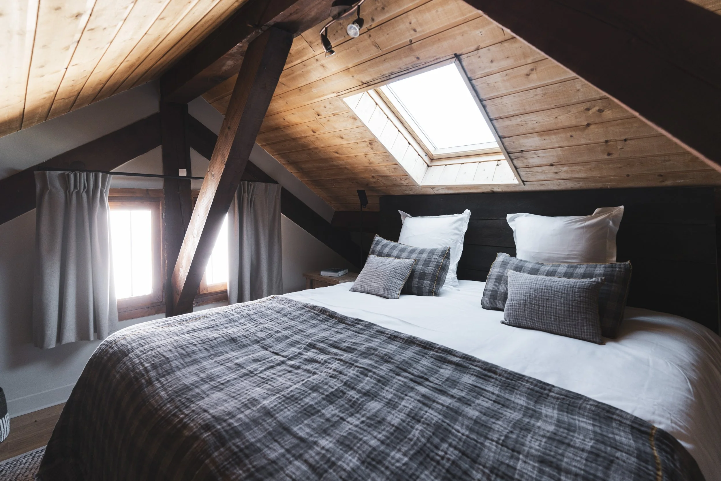 Cozy bedroom in an attic with a sloped wood-paneled ceiling, skylight, and small window with curtains. The bed has white pillows and gray plaid bedding, with a wooden nightstand on one side.