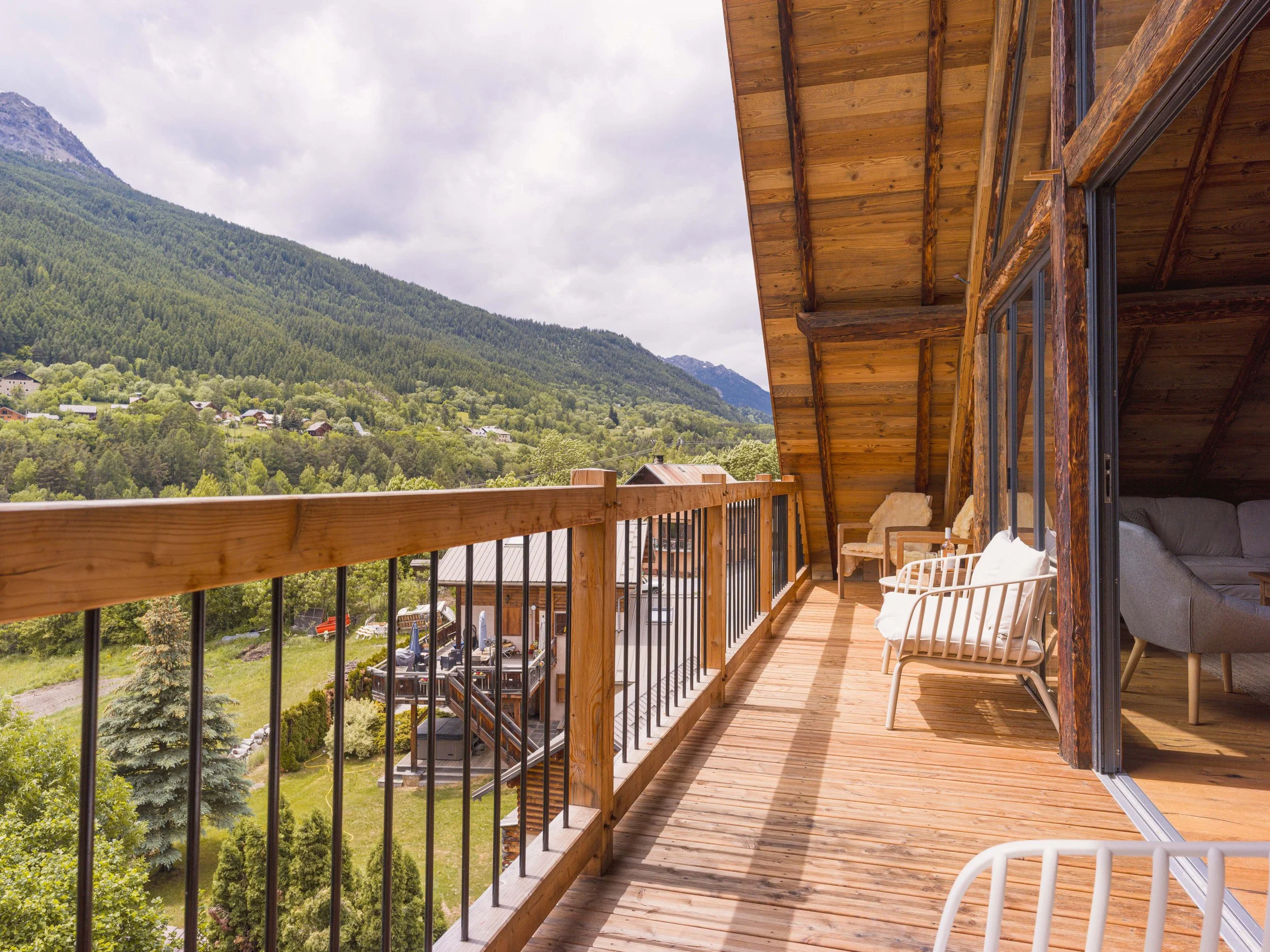Vue depuis un balcon sur un paysage montagneux boisé sous un ciel nuageux, avec un parquet, une balustrade et du mobilier d'extérieur, notamment des chaises.
