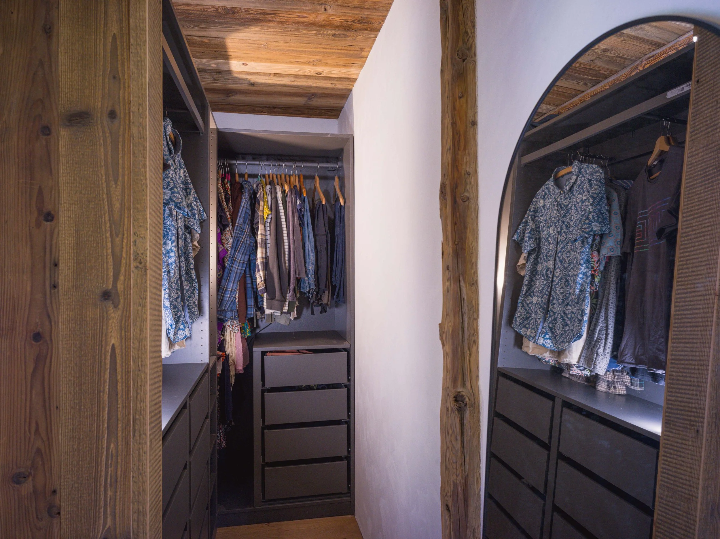 Un dressing avec des vêtements suspendus à des tringles métalliques et des étagères de chaque côté, encadré par des poutres en bois et un plafond en bois.