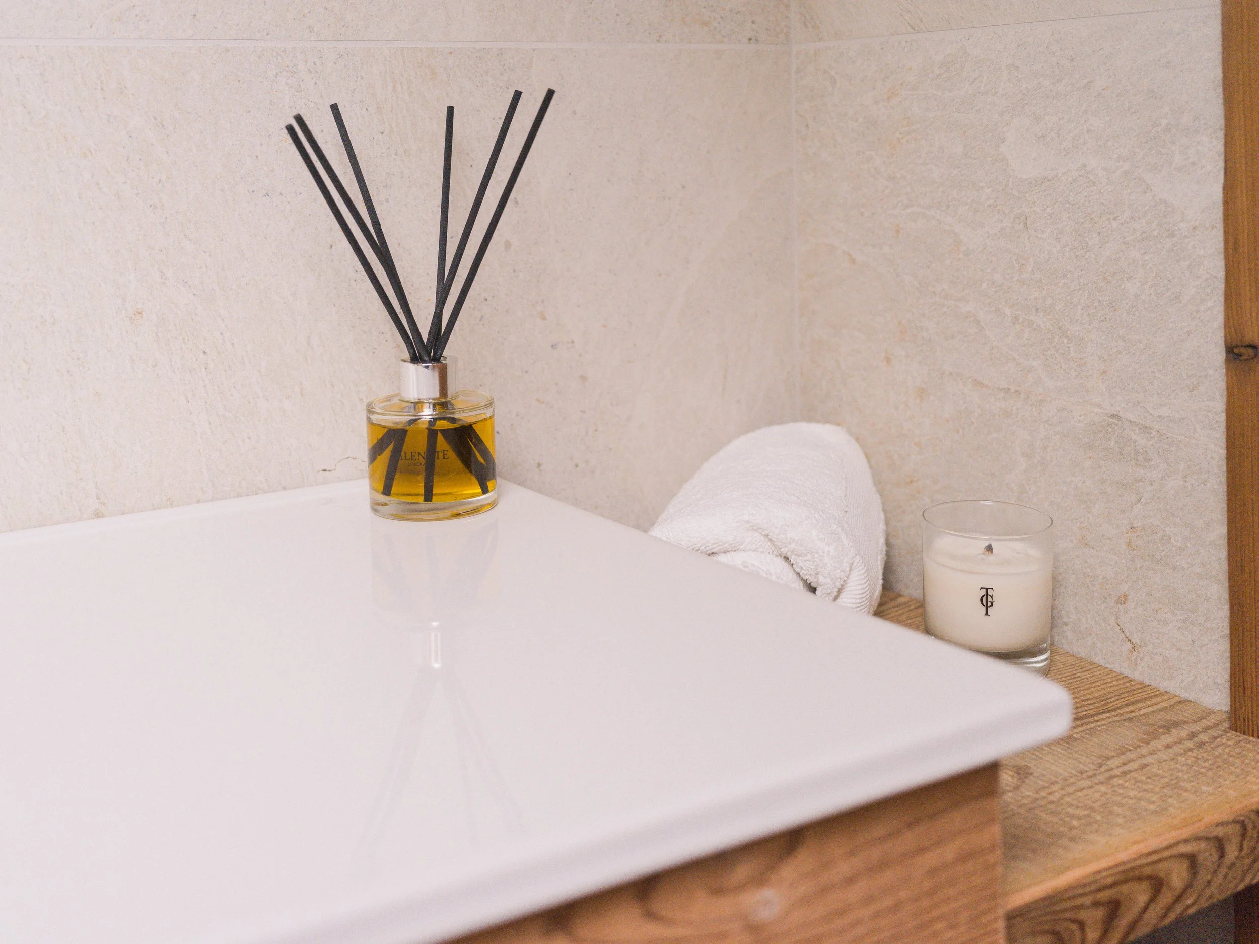Un plan de toilette sur lequel sont posés un diffuseur à bâtonnets, une serviette blanche enroulée, une bougie et un récipient en verre, avec des murs carrelés de couleur beige et des touches de bois.