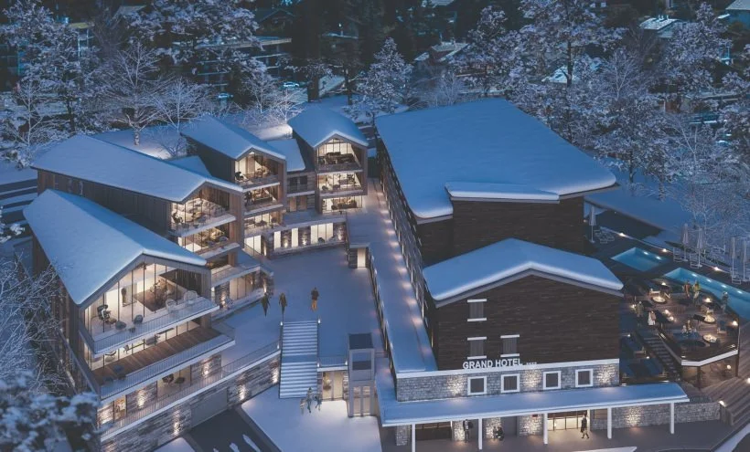 Aerial night view of a snow-covered hotel complex with multiple buildings and illuminated outdoor areas surrounded by snowy trees.