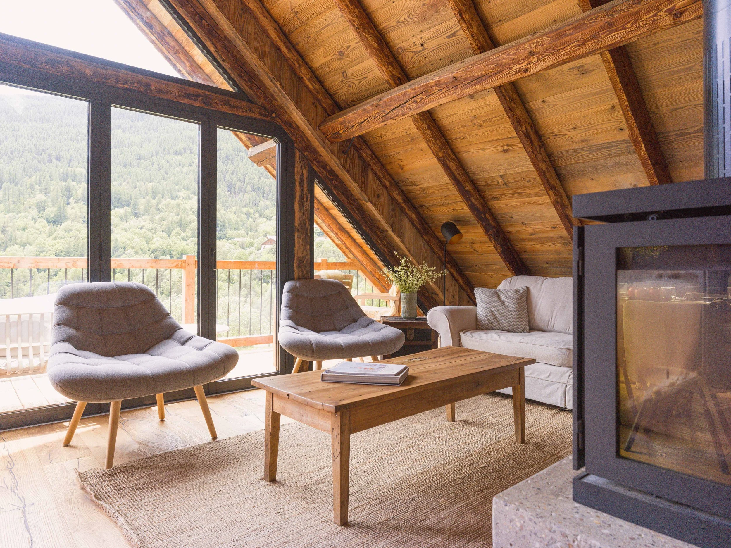 Salon doté de grandes baies vitrées offrant une vue sur la montagne, d'un plafond en bois avec des poutres apparentes, d'un canapé beige orné de coussins, d'une table basse en bois sur laquelle sont posés des livres, d'une plante sur une table d'appoint et d'une cheminée.