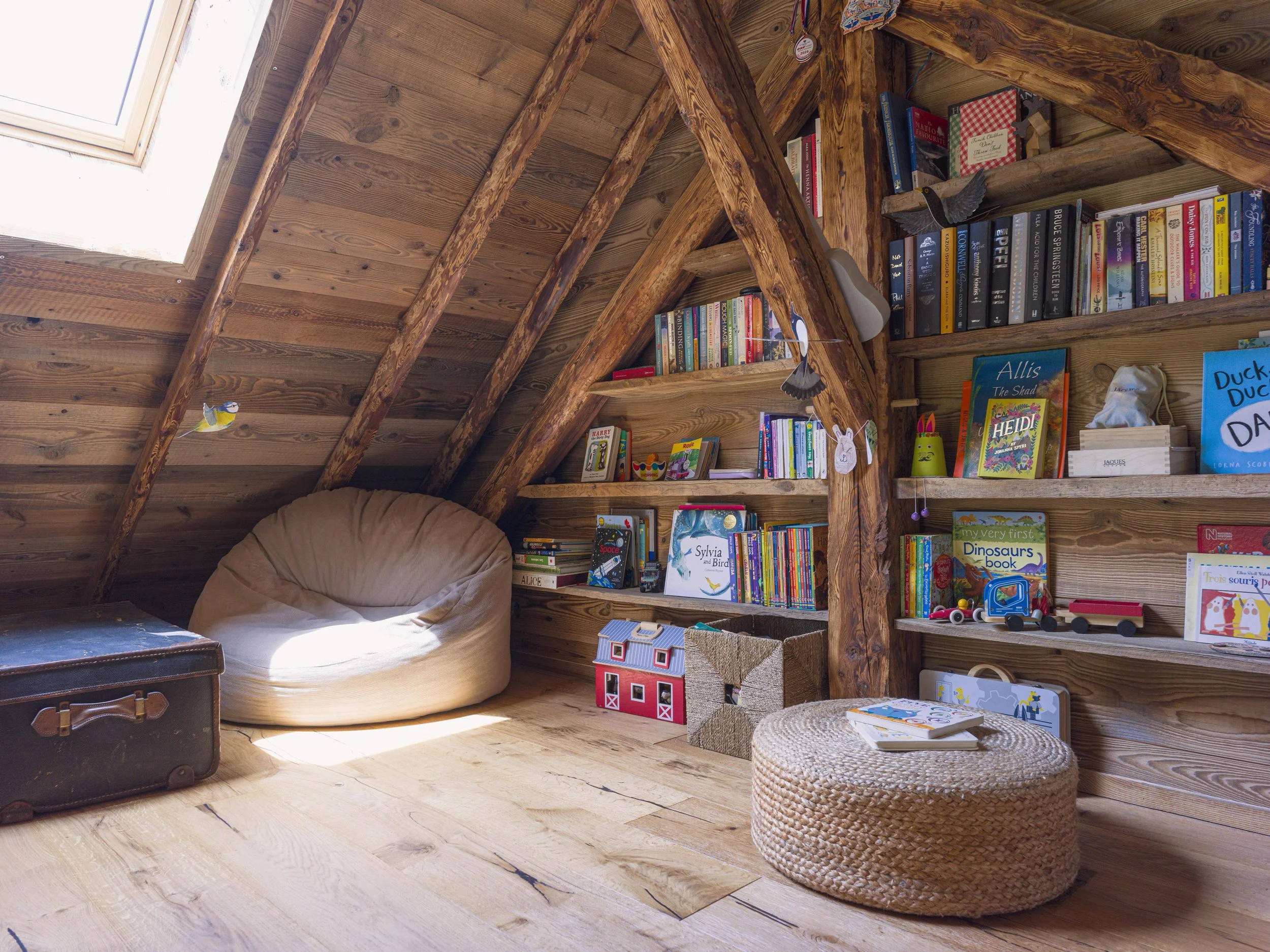 Un grenier chaleureux aux murs et au plafond en bois, doté d'une lucarne, d'étagères encastrées remplies de livres pour enfants, d'un grand pouf beige, d'un tabouret rond tressé, d'une valise vintage et d'objets décoratifs tels que des découpages en papier représentant des animaux