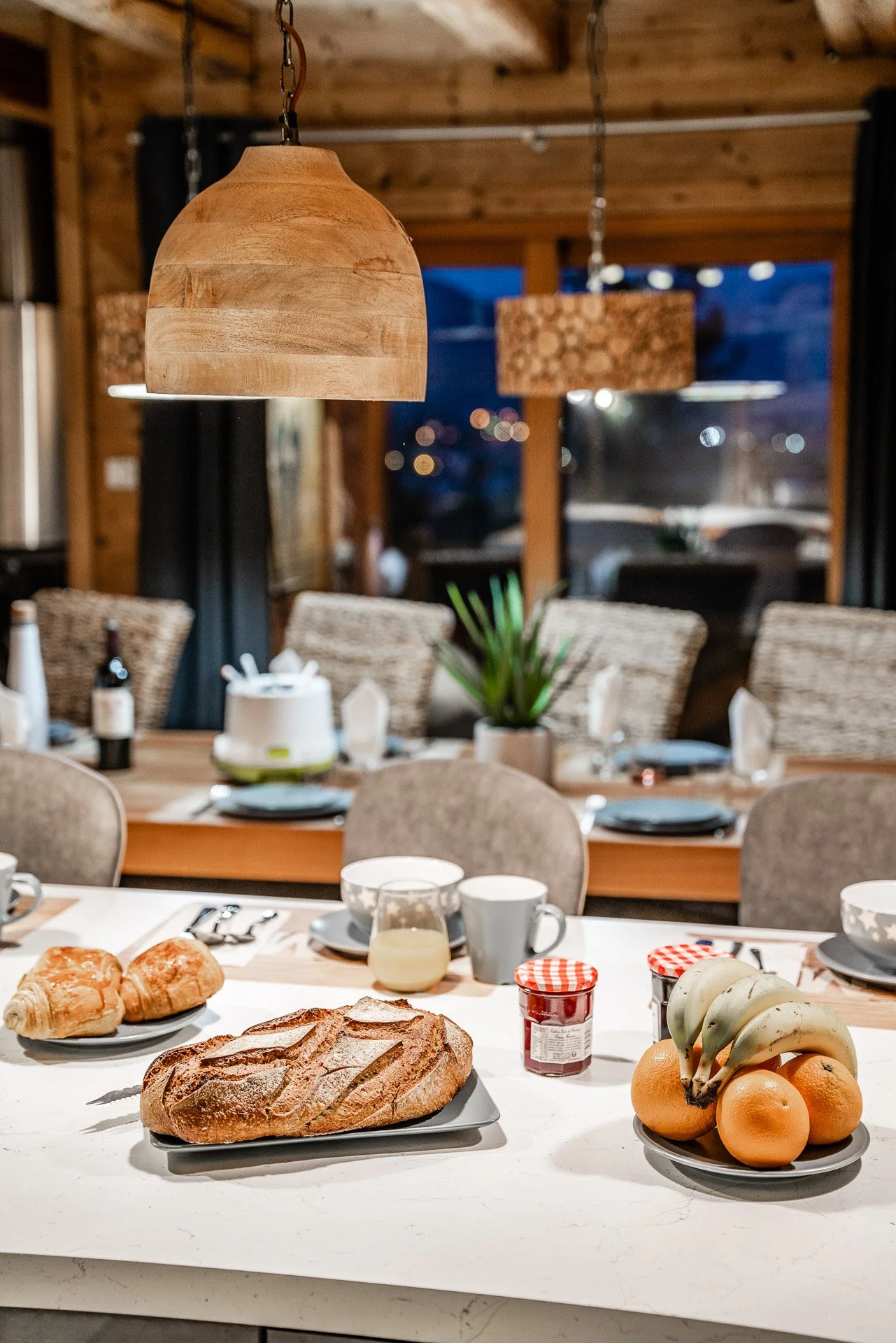 Breakfast table with croissants, bread, bananas, oranges, jelly, mug, glass of orange juice, and plates set for a meal in a cozy wooden dining room with pendant lights and window view.