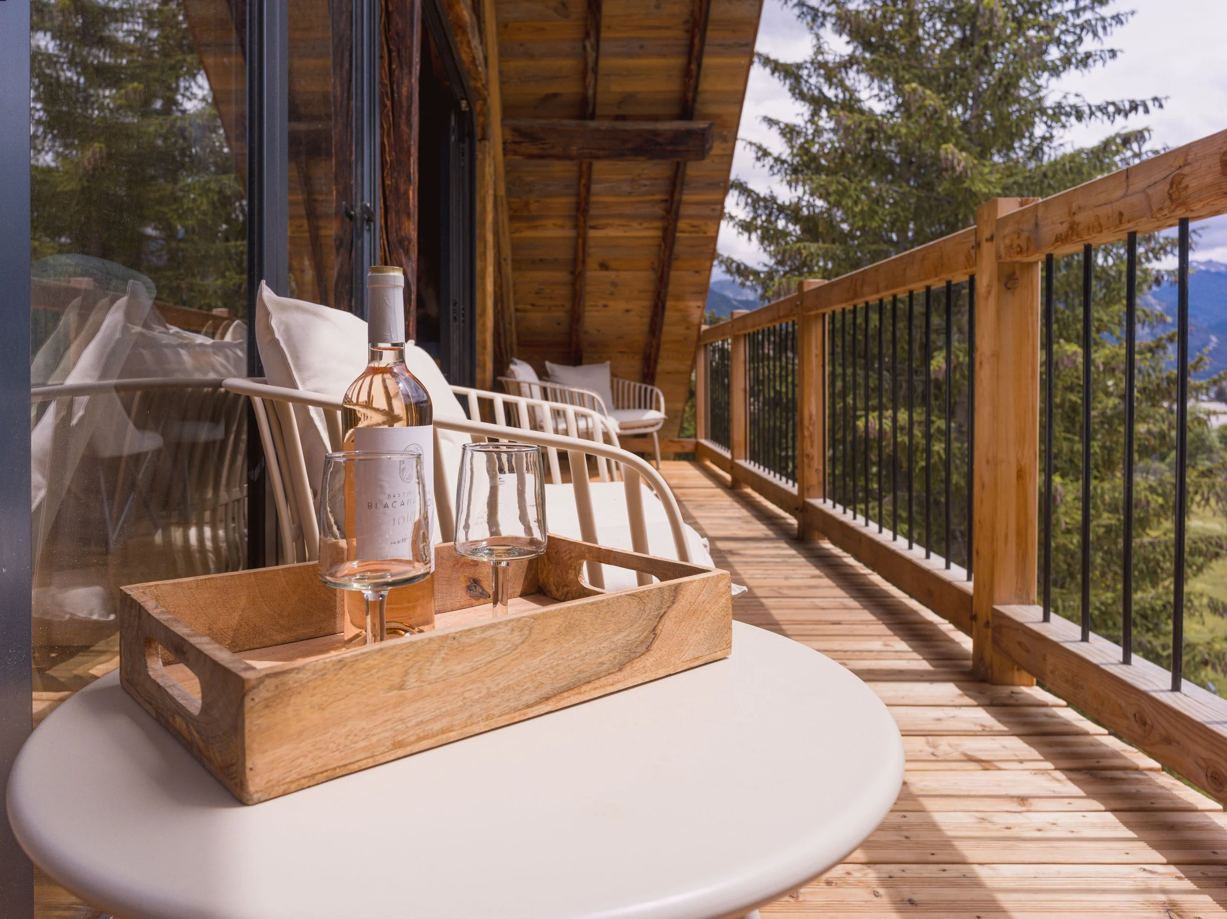 Balcon extérieur avec balustrade en bois, chaises rembourrées et une table blanche dressée avec une bouteille de vin, des verres et un plateau en bois, offrant une vue imprenable sur les montagnes.
