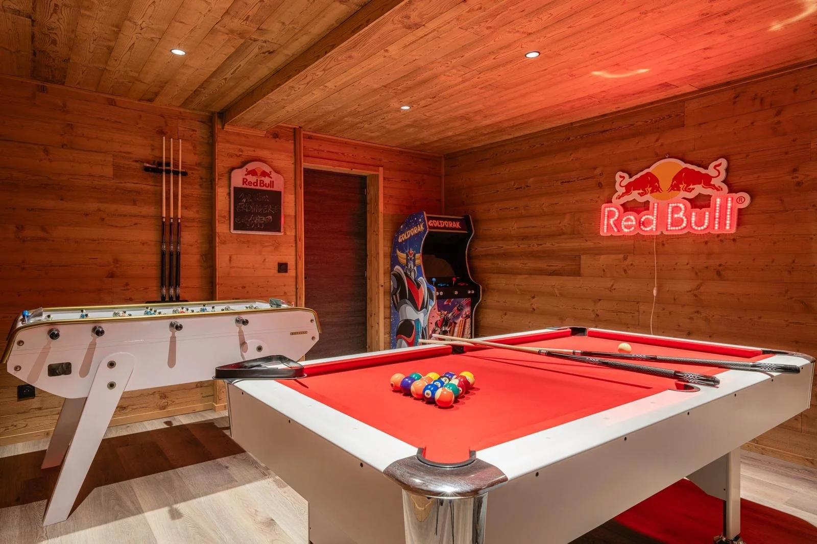 Wood-paneled game room with a red-felt pool table, billiard balls racked, two pool cues, a white air hockey table, a Golden Drak arcade machine, and Red Bull neon sign on the wall.