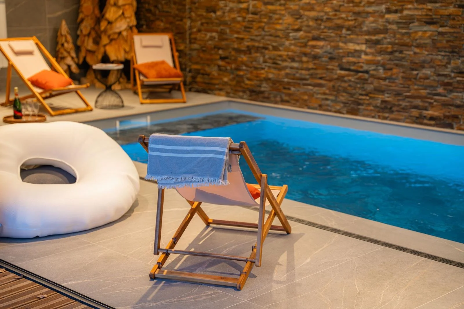 Indoor swimming pool area with a wooden lounge chair draped with a blue towel, a large inflatable white pool float, several wooden lounge chairs with orange cushions, a small table with a bottle of champagne and glasses, and a stack of firewood again