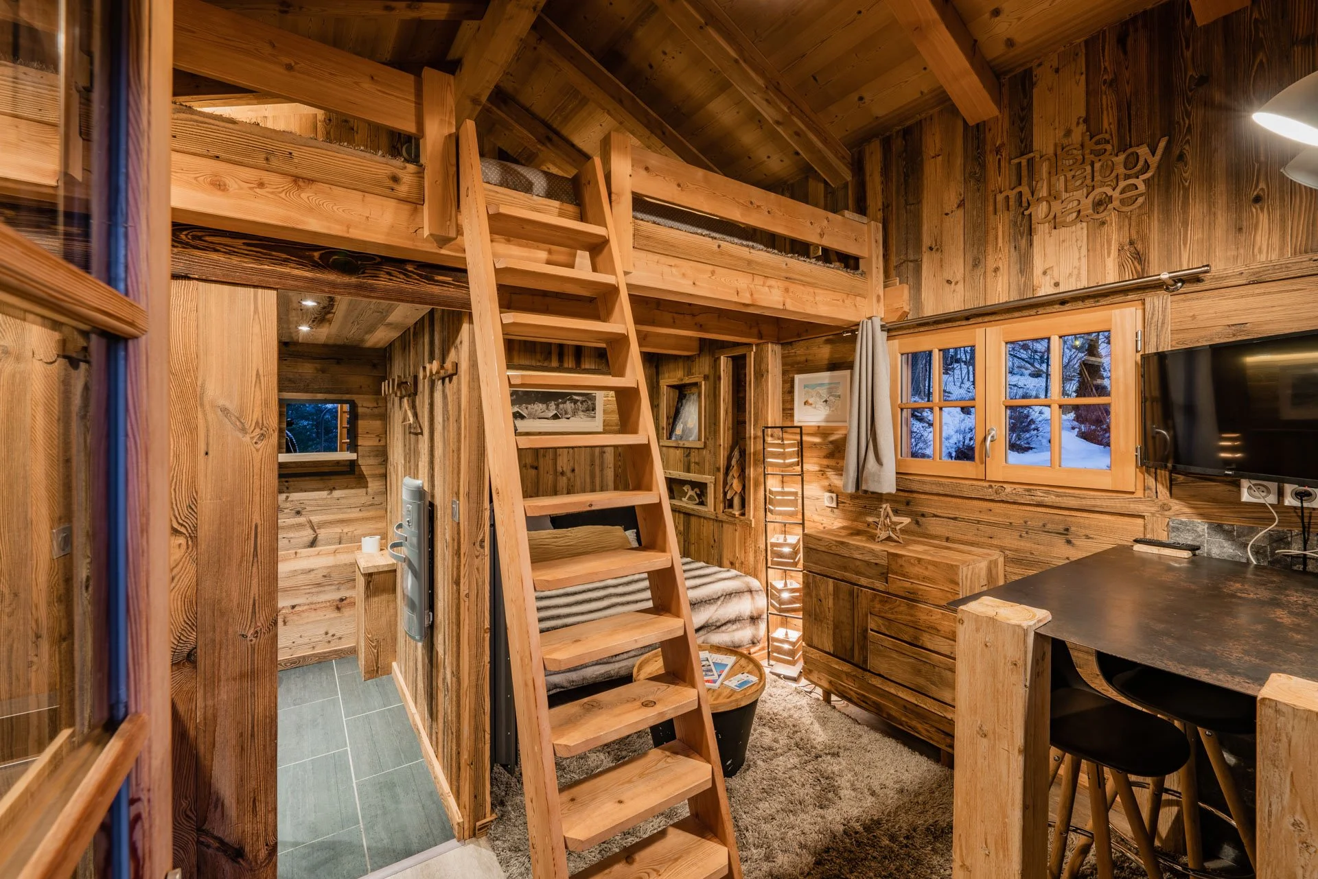 Cozy wooden cabin interior with loft area, small bed, window showing snow outside, flat-screen TV, and rustic decorations.