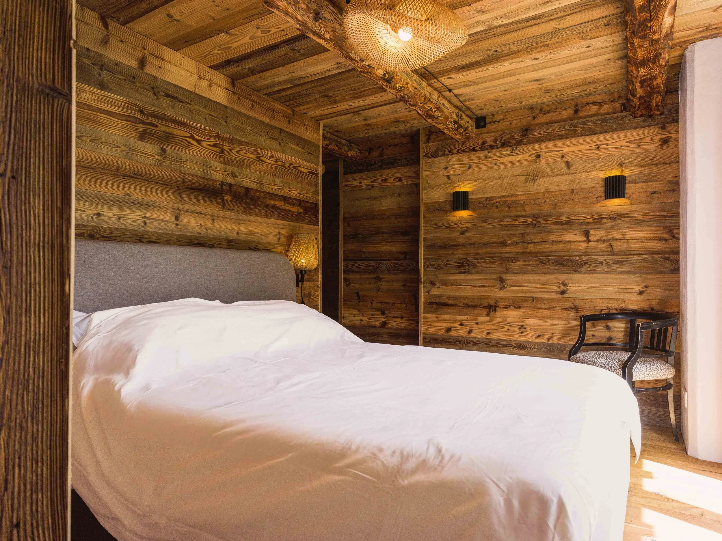 Une chambre aux murs, au plafond et aux poutres en bois, avec un lit aux draps blancs, une petite lampe posée sur une table de chevet et une chaise noire dotée d'un coussin à motifs.