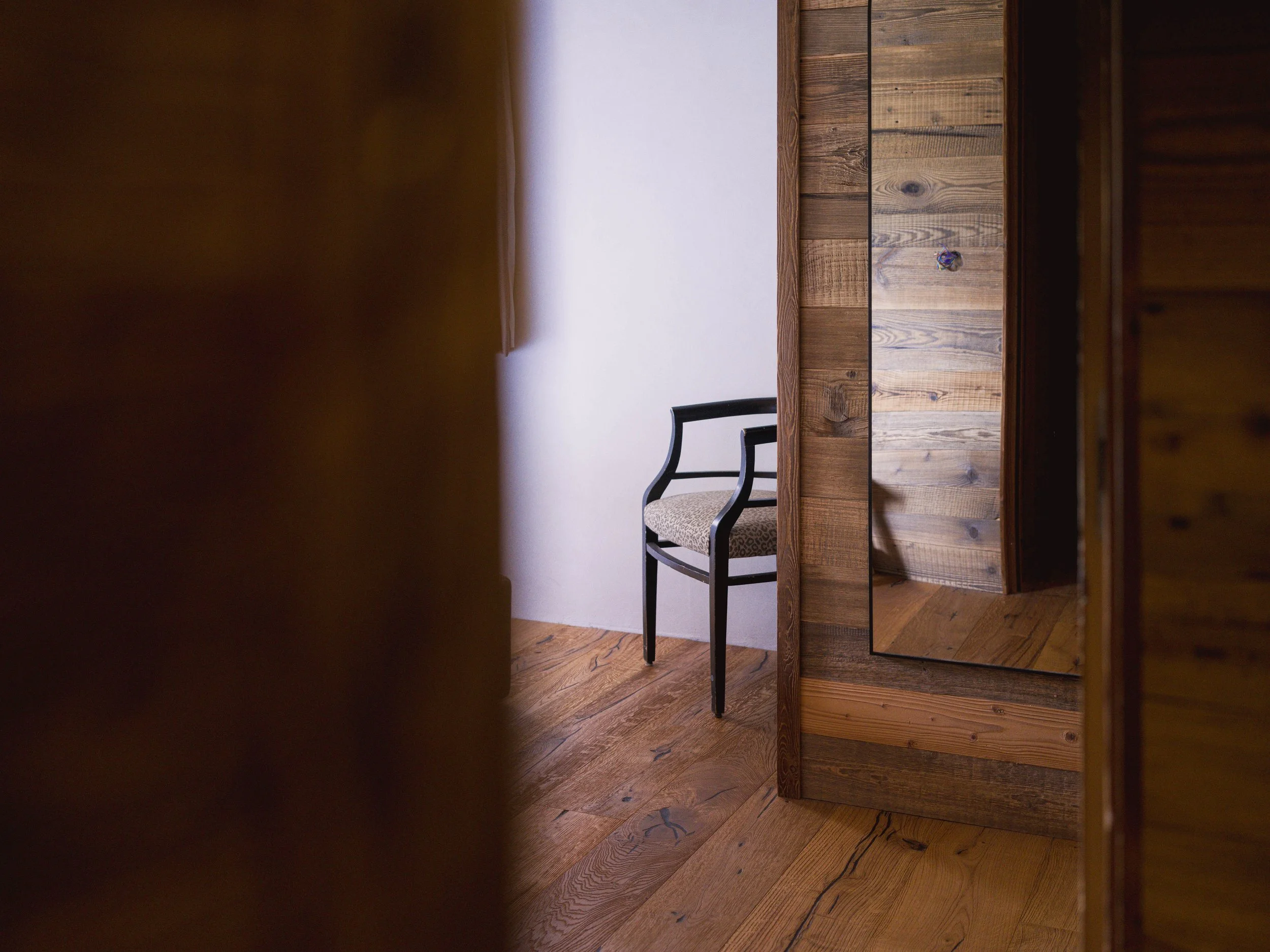 Une partie d'une pièce où l'on voit une table en bois, une chaise rembourrée à la structure noire et un mur blanc. La photo a été prise à travers une fente ou une ouverture, ce qui crée une bordure sombre à gauche et à droite.