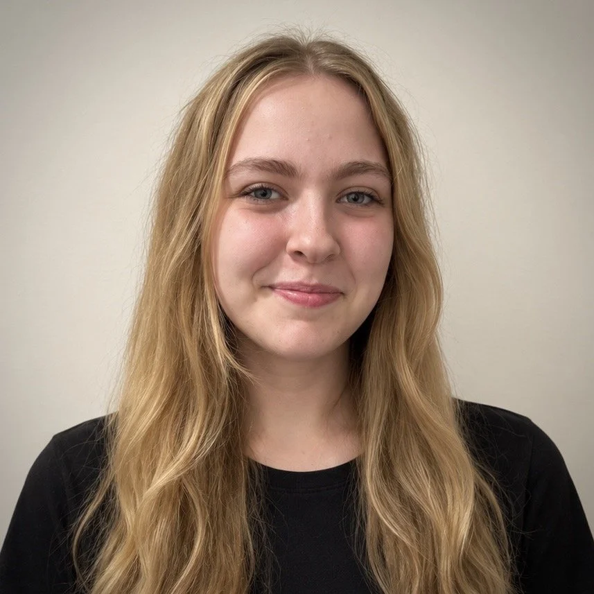 A young woman with wavy brown hair and blue eyes, wearing a black top, smiling softly at the camera against a plain light-colored background at cherubsnurseryschool.com