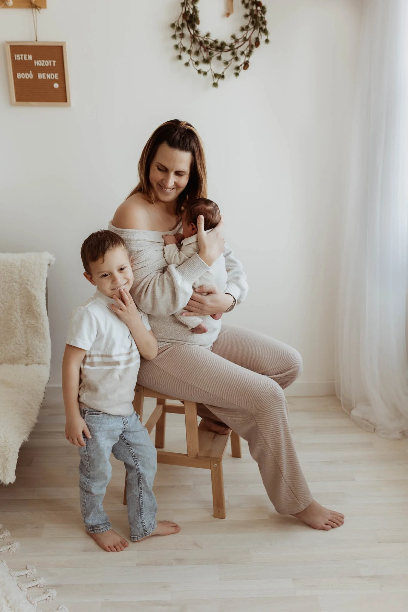 A woman sitting on a wooden chair, smiling and holding a baby in her arms. A young boy is standing next to her, touching his face, in a cozy, well-lit room with white walls and curtains.