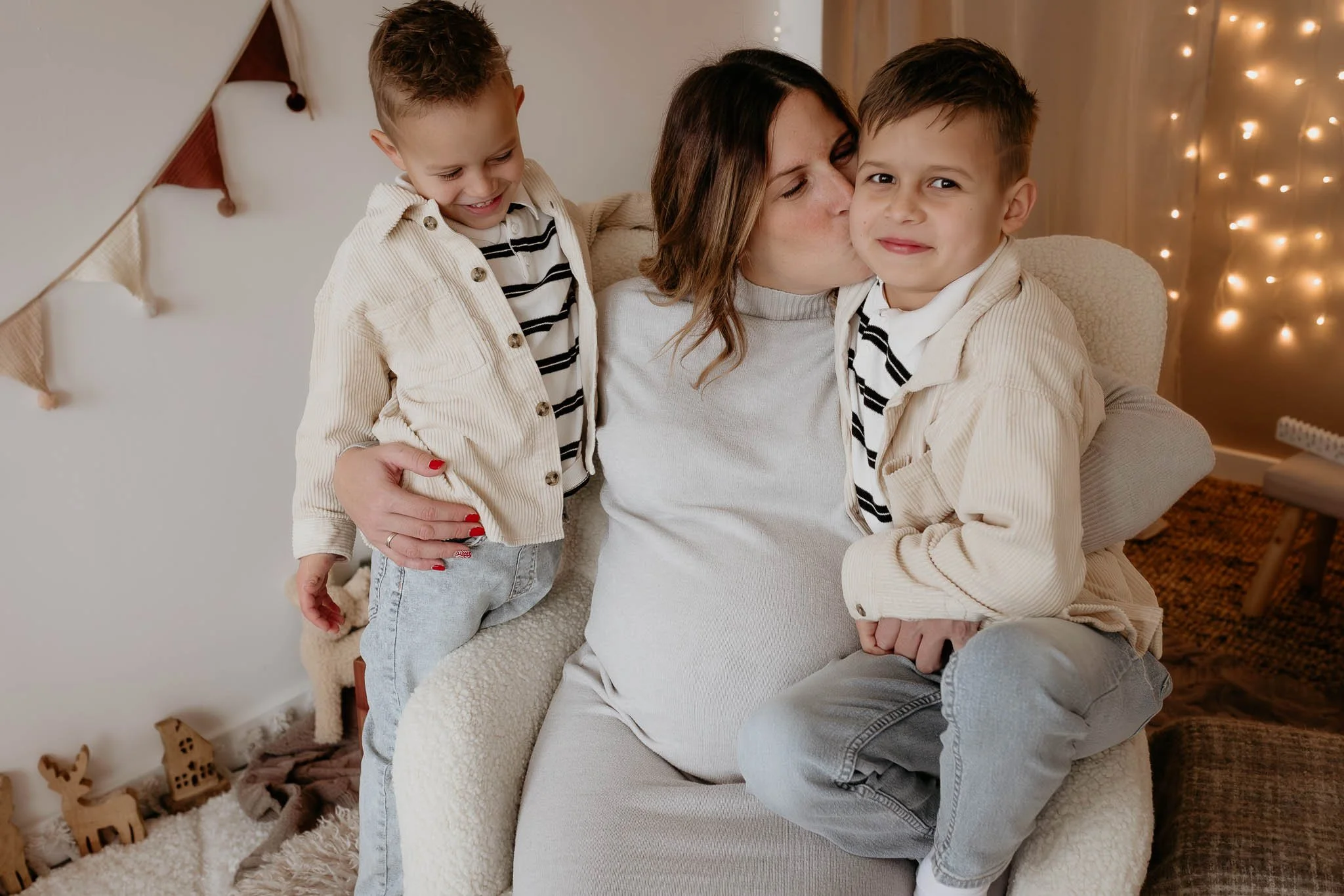 A woman with two young boys sitting on a cozy armchair, one boy is giving her a kiss on the cheek, while the other boy is smiling. The scene is decorated with warm fairy lights and holiday decorations in the background.