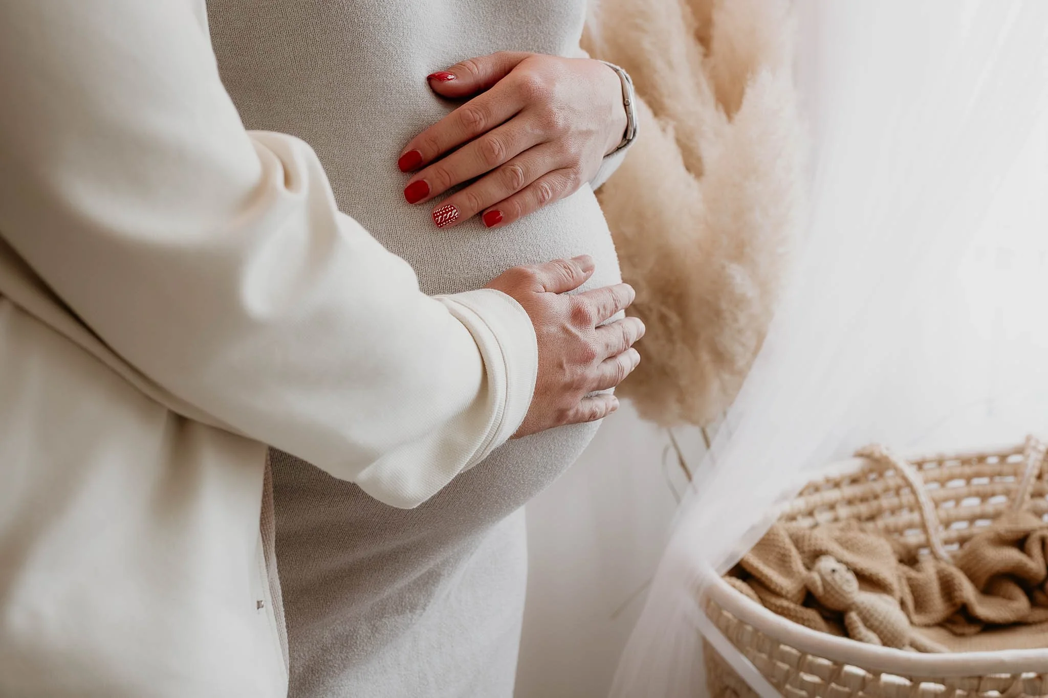 Close-up of a pregnant woman and partner holding her belly. The woman has red manicure with a patterned accent nail. A laundry basket with clothes is visible in the background.