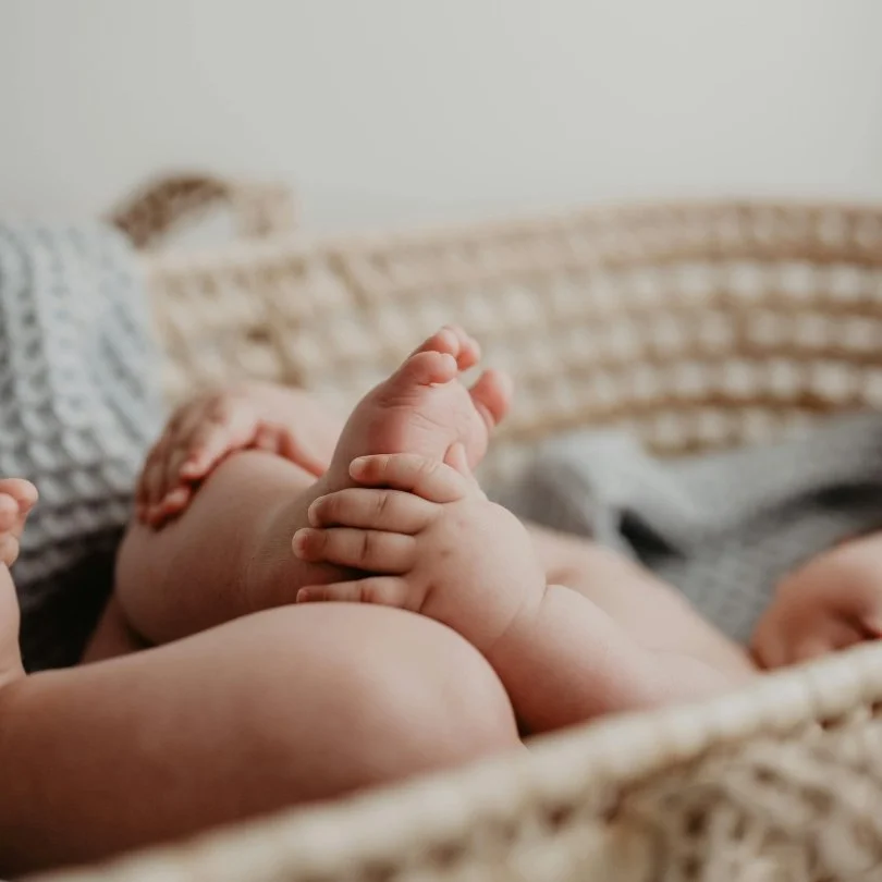 Close-up of a baby's legs and hands, lying in a woven basket on a bed with cushions in the background.