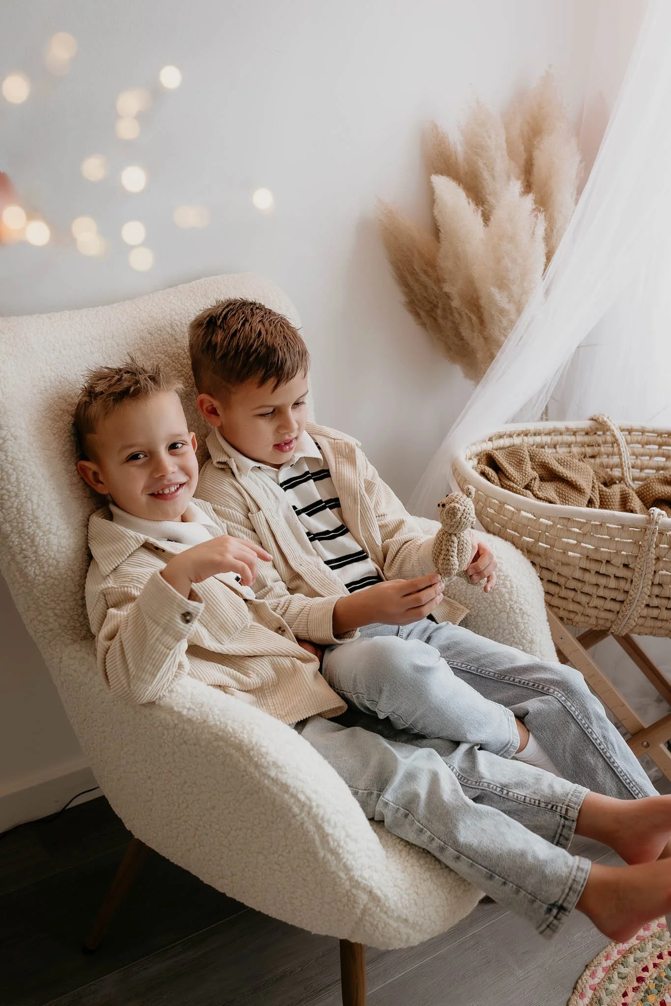 Two young boys sitting on a cozy, cream-colored armchair in a warmly decorated room, with one boy smiling at the camera and the other playing with a small crochet teddy bear, next to a wicker bassinet and tall pampas grass decor.