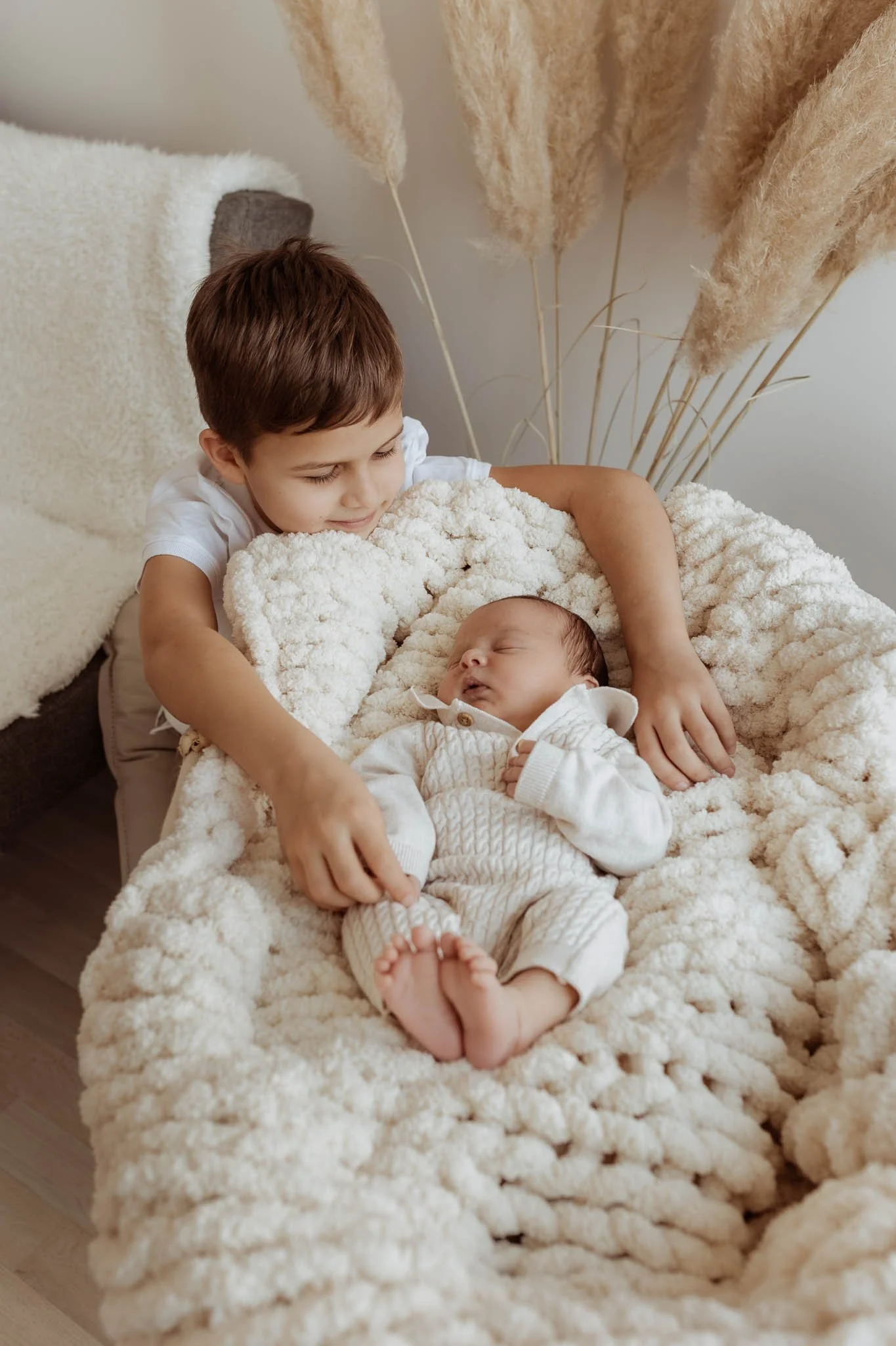 A young boy lovingly holding a sleeping baby on a cozy, fluffy blanket in a warm setting.