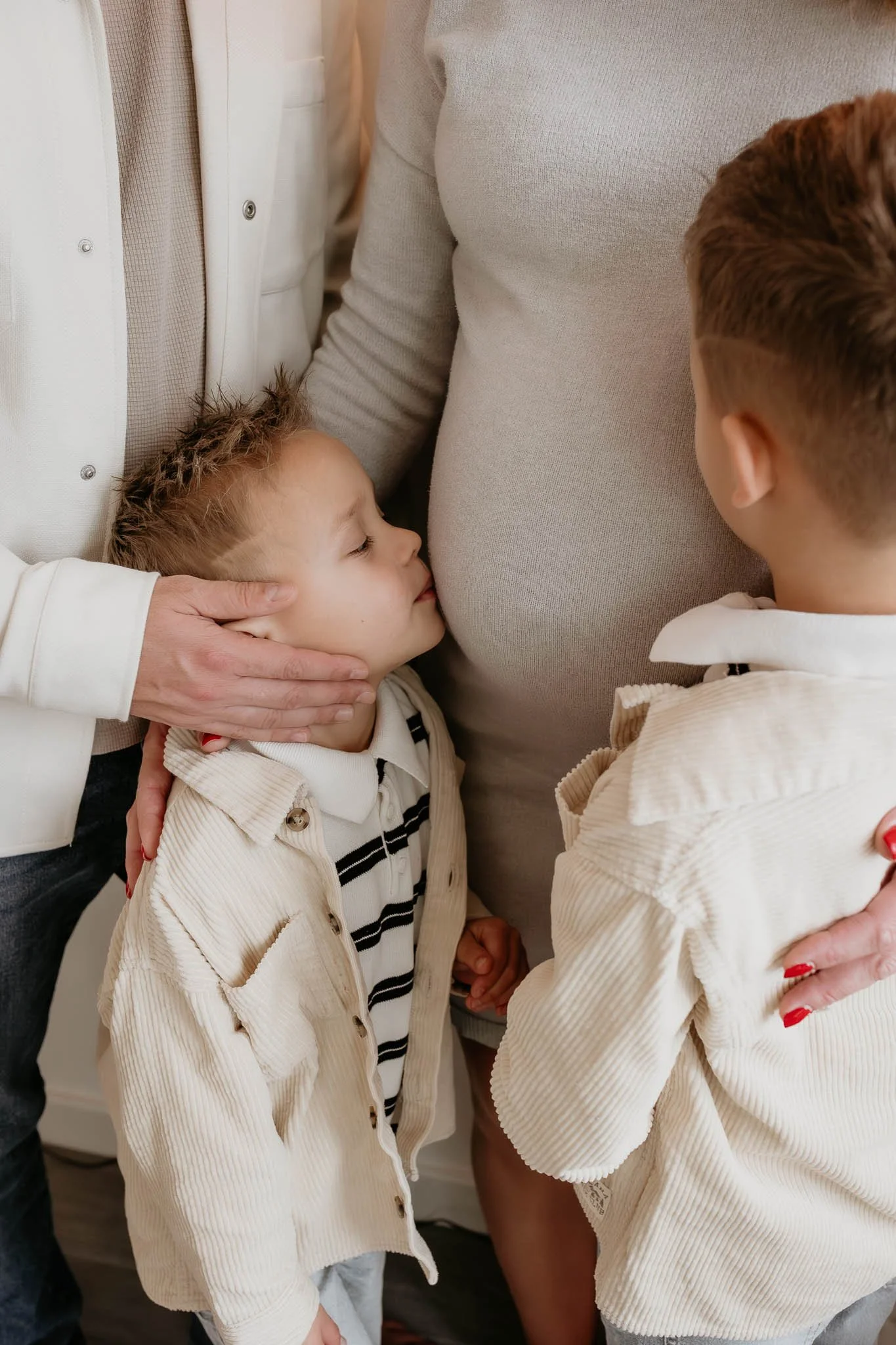 A young boy with closed eyes leaning his head against a pregnant woman's belly, being gently touched on the forehead by another adult. The woman is wearing a light-colored top, and the boy and another child are dressed in light-colored clothing. Hand