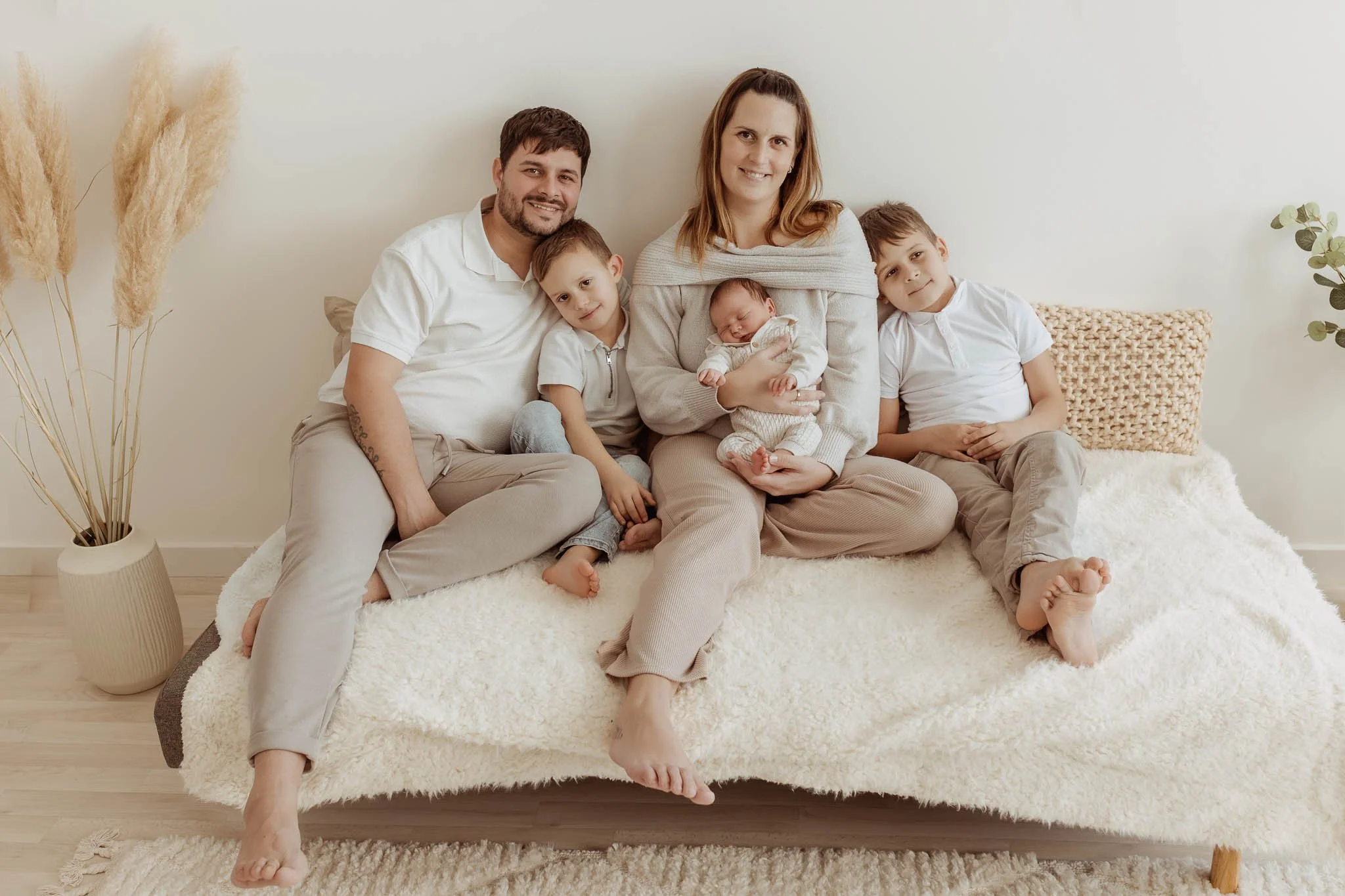 Family of five sitting on a cozy bed in a minimalist, neutral-colored room, with dried pampas grass and a woven pillow visible.