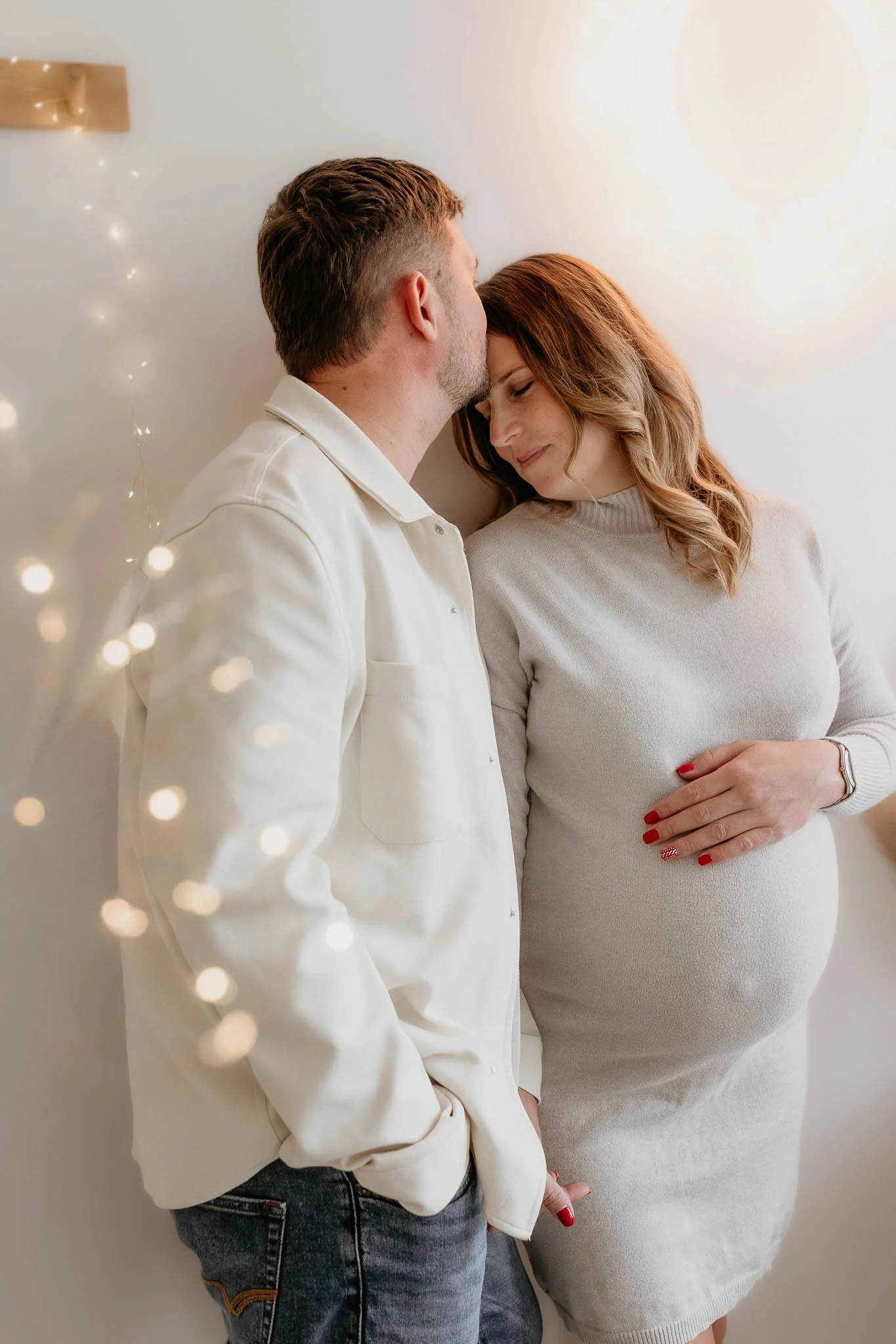A man and a pregnant woman standing close together indoors, with soft lighting and fairy lights in the foreground. The woman is touching her belly and smiling with her eyes closed, while the man is kissing her forehead.