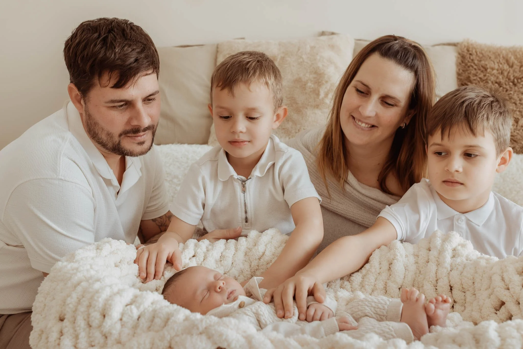 Family of five with two young boys, a man, a woman, and a newborn baby lying on a bed surrounded by soft blankets, gently touching the sleeping baby.