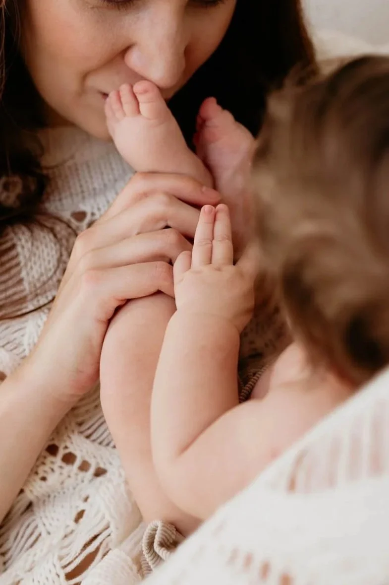 A woman kissing a baby's foot while holding their leg, with both of them lying close together.