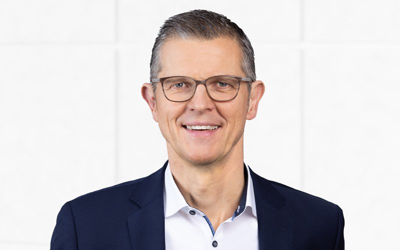 Professional man in glasses smiling, wearing a dark blazer and white shirt, standing against a plain light background.