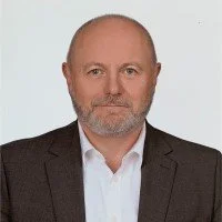 A portrait of a middle-aged man with a beard and short hair, dressed in a dark blazer and light shirt, against a plain light background.