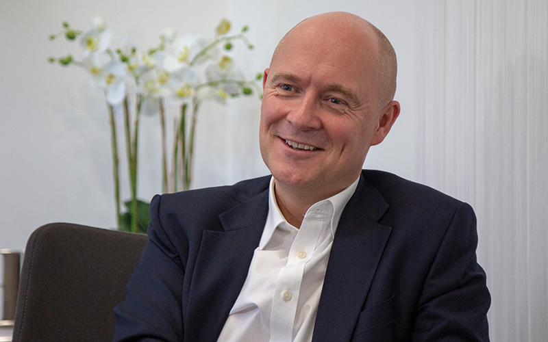 A smiling man in a suit sitting in an office with white walls and a vase of white orchids in the background.