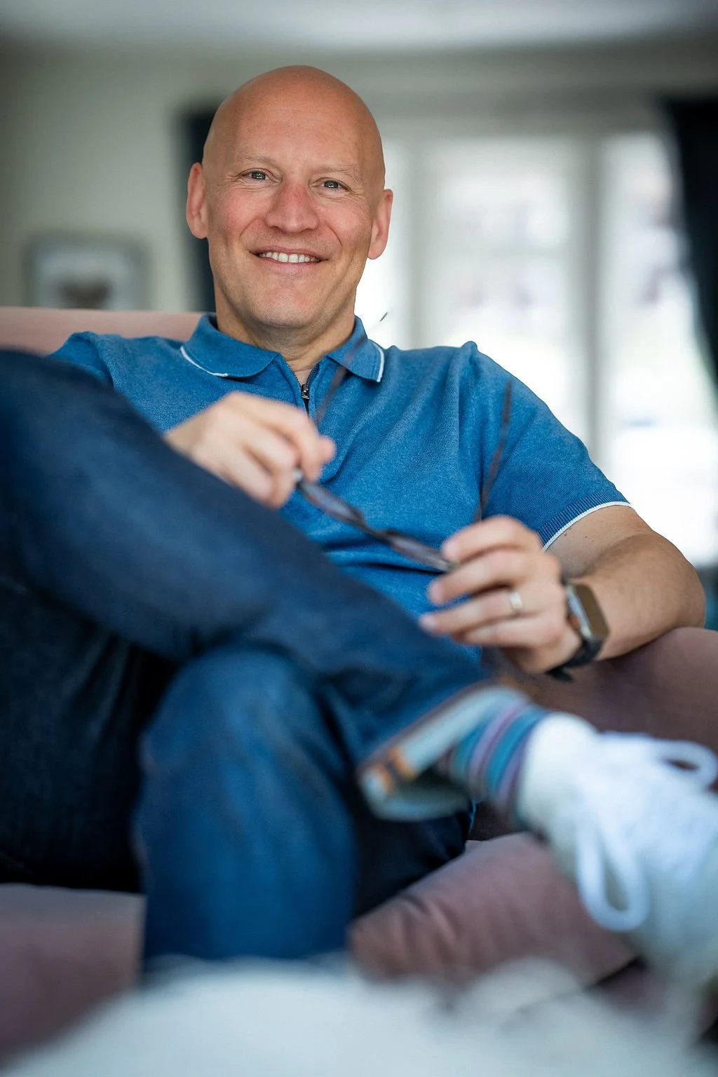 A smiling man with a bald head sitting on a couch in a bright room, wearing a blue polo shirt, striped socks, and a watch, holding sunglasses.