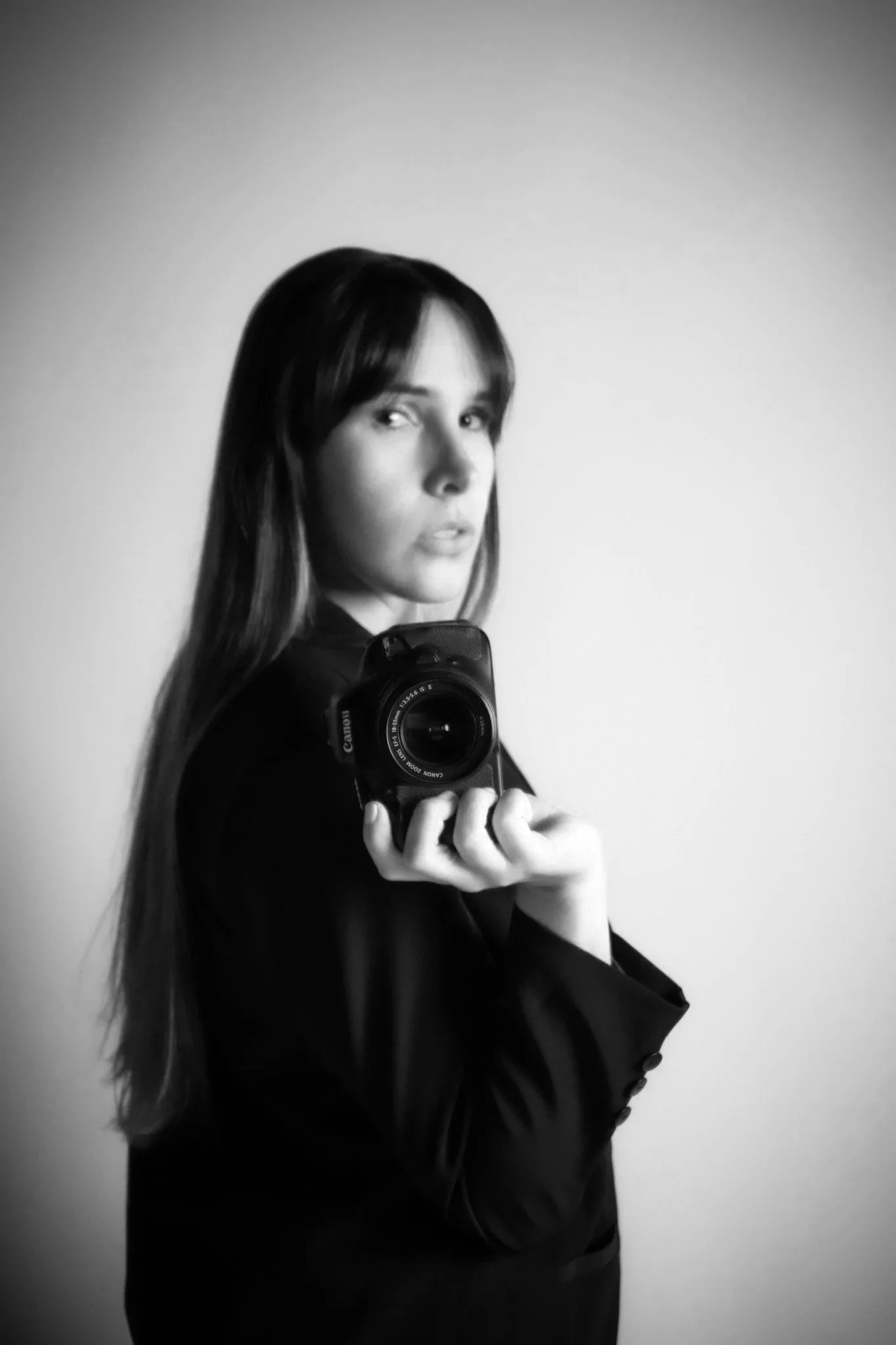 A woman taking a mirror selfie with a camera in her hand, wearing a black blazer, against a plain light gray background.