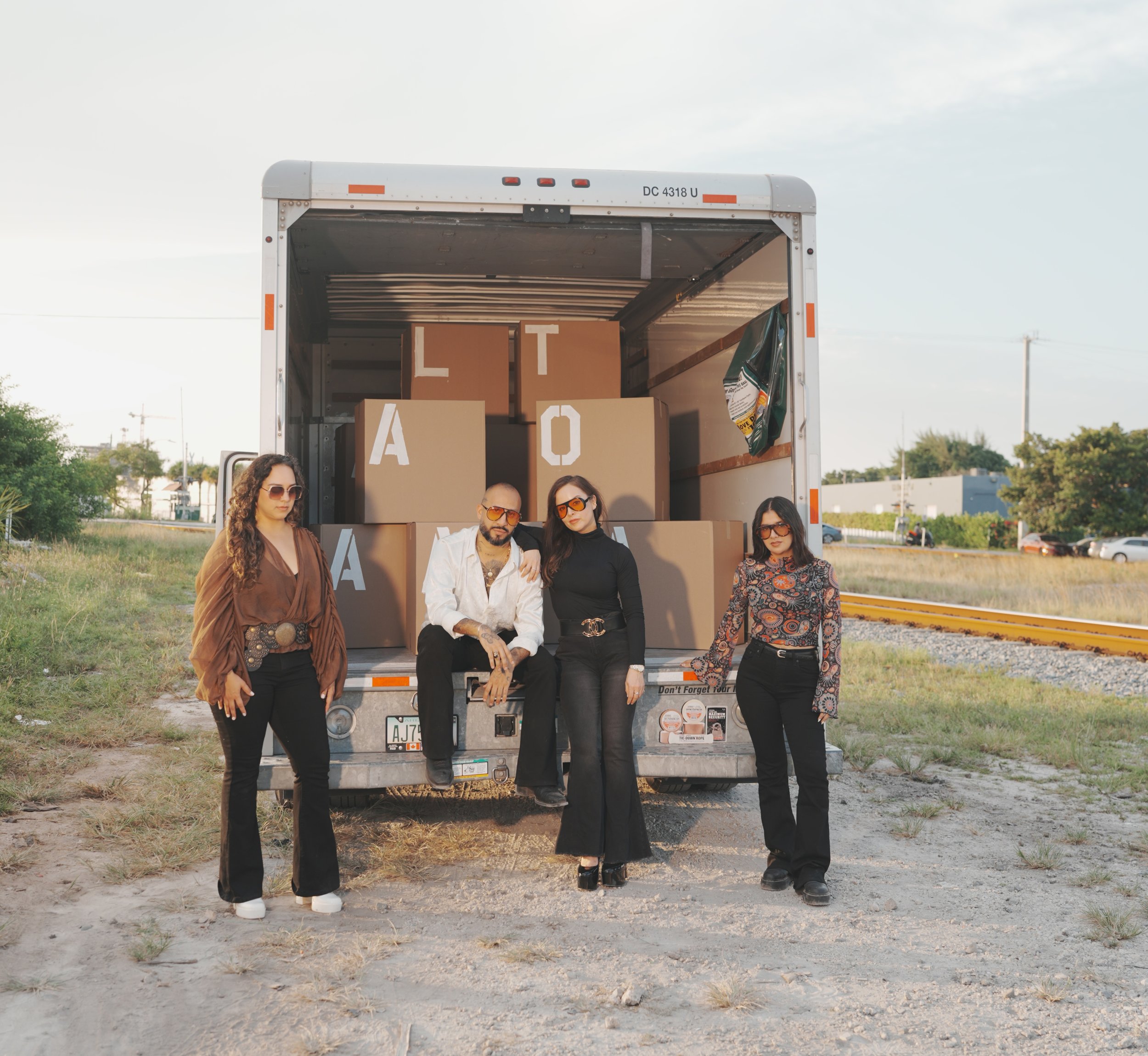 A group of five diverse young adults posing in front of an open moving truck loaded with cardboard boxes, outdoors during daytime with a cloudy sky.