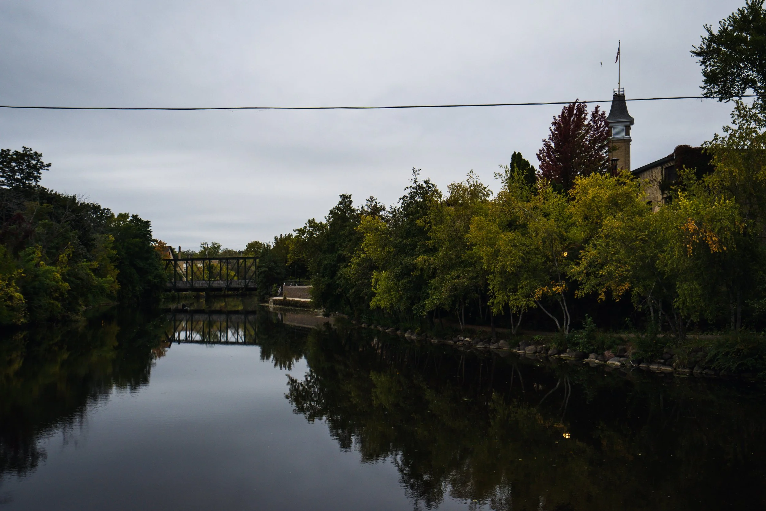 A peaceful river reflecting overhanging trees and a church steeple under an overcast sky.