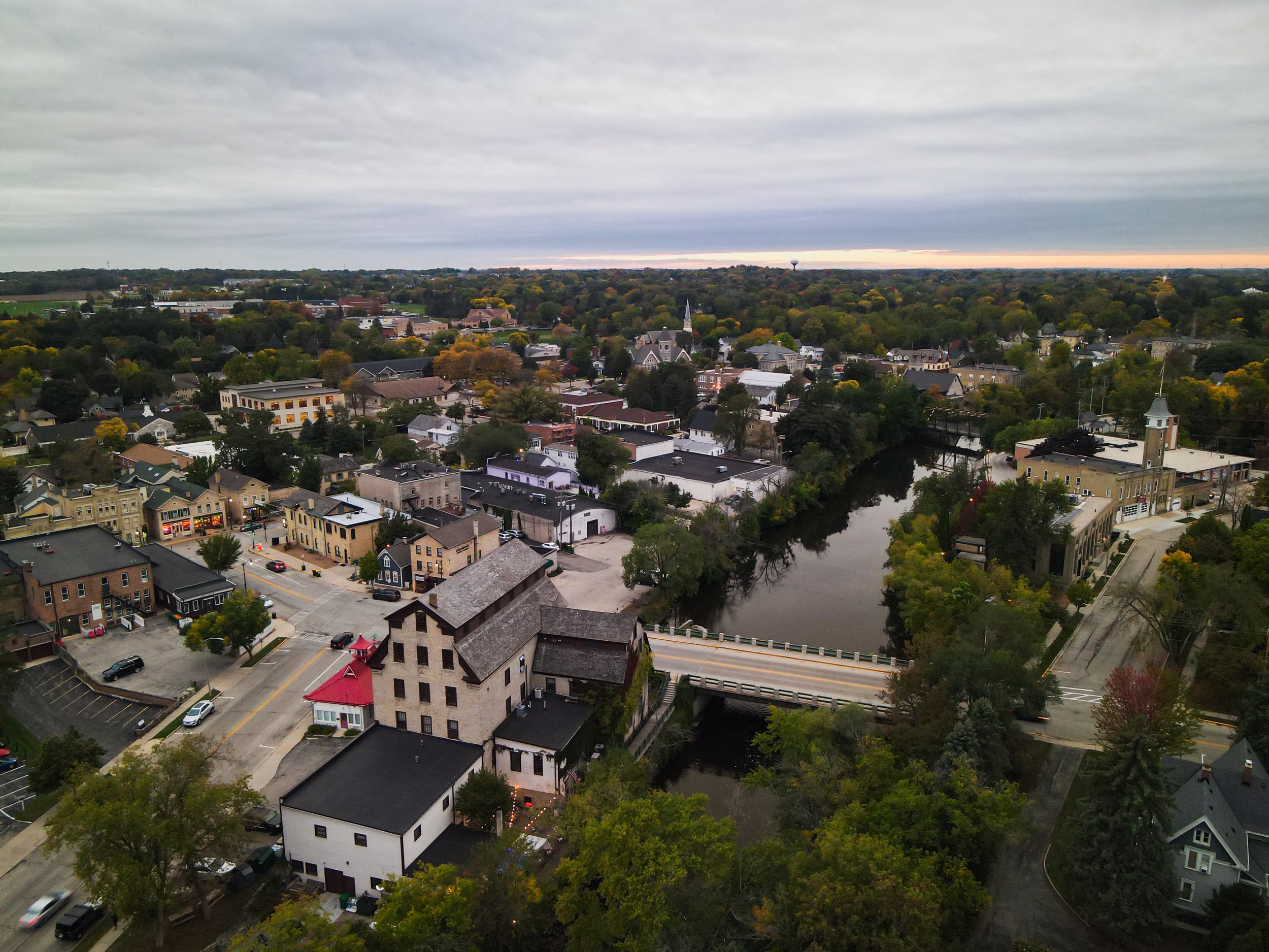 Aerial view of a small town with houses, trees, a river, a church, and a water tower under a cloudy sky during twilight.