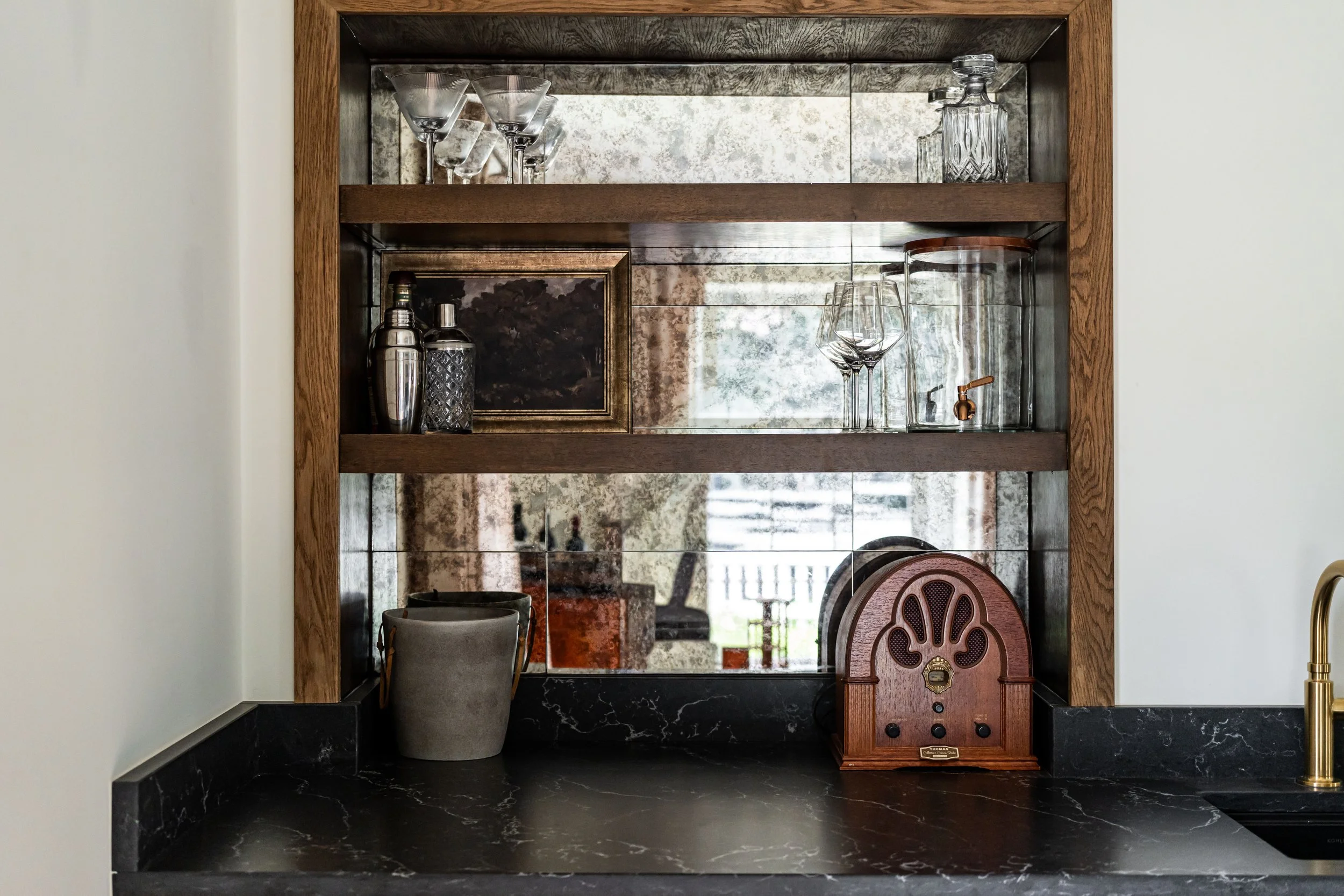 A wooden and mirrored bar shelf holding glassware and bottles, with a vintage radio and a cup on a black marble counter below, and a reflection of a window outside.