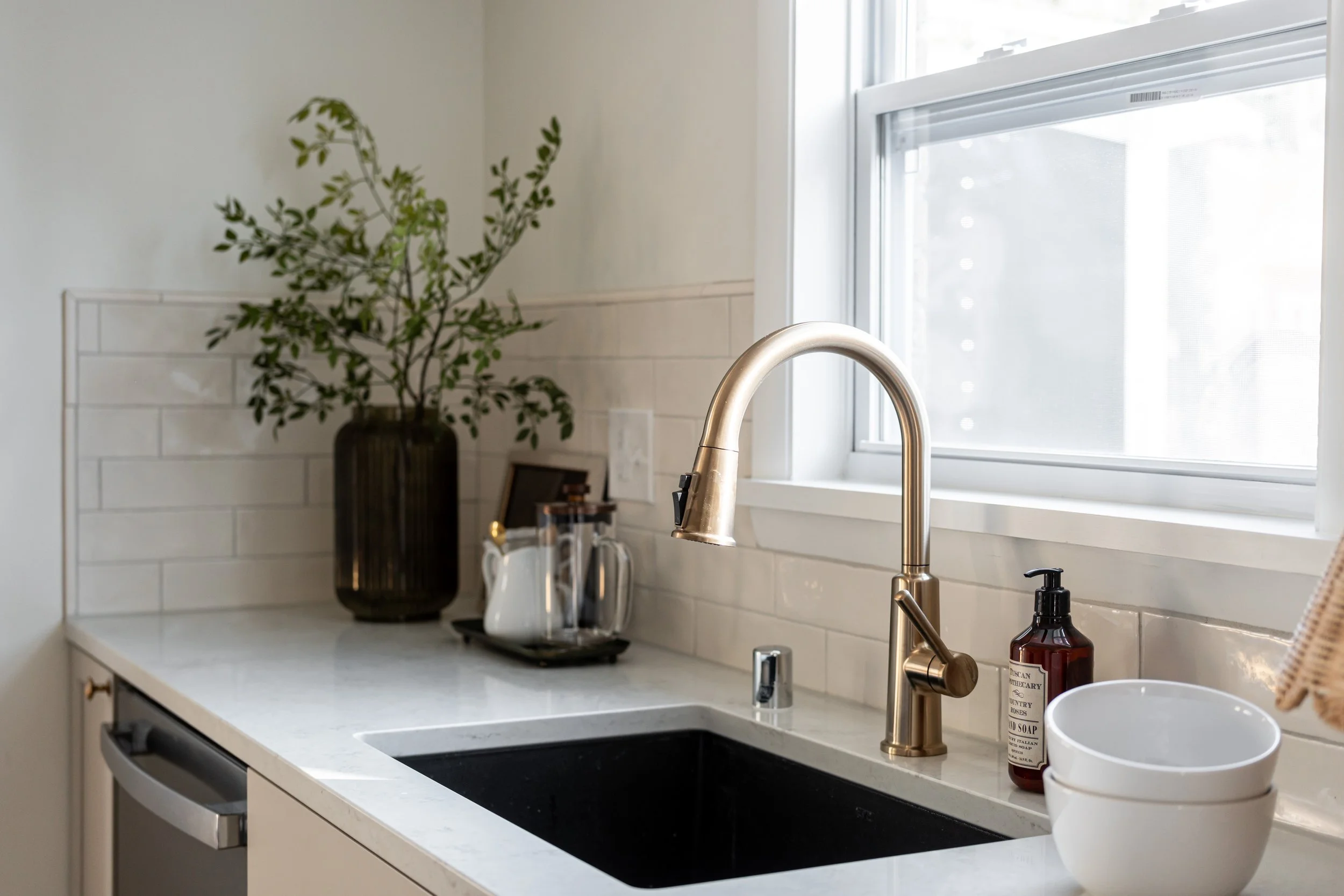 Kitchen sink with a gold faucet, white countertop, a potted plant in a dark vase, soap dispenser, mugs, and a window letting in natural light.