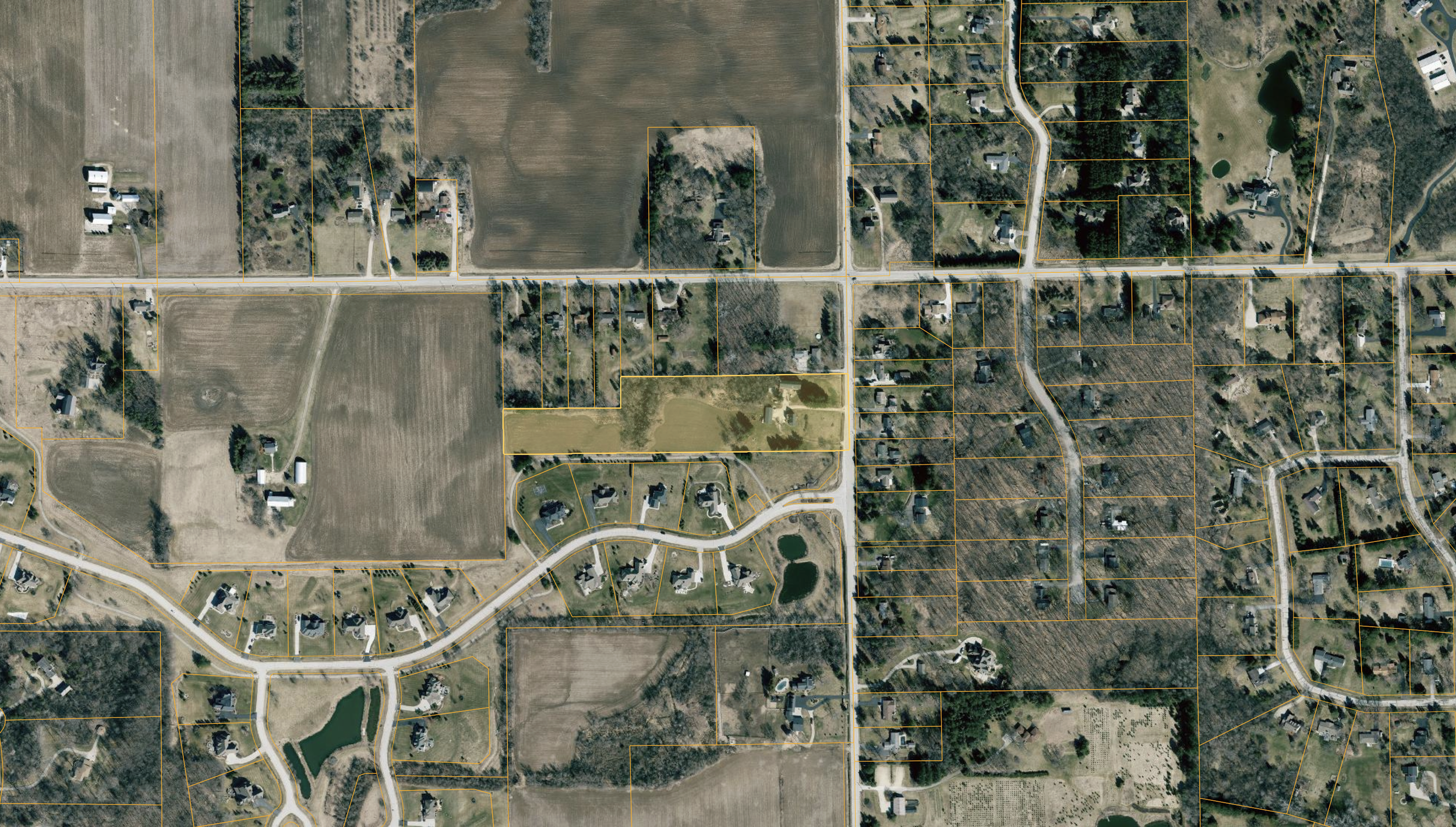 An aerial view of a suburban area showing houses, roads, trees, and open fields, with property boundaries marked by orange lines.