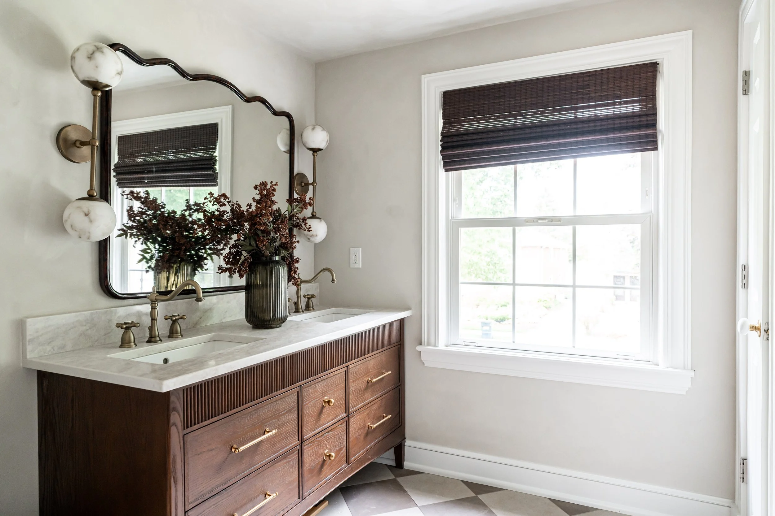 Bathroom vanity with a dark wood cabinet, light-colored marble countertop, gold hardware, and a large mirror illuminated by wall-mounted globe sconces. There is a vase with flowers on the counter, and a window with brown venetian blinds letting in na