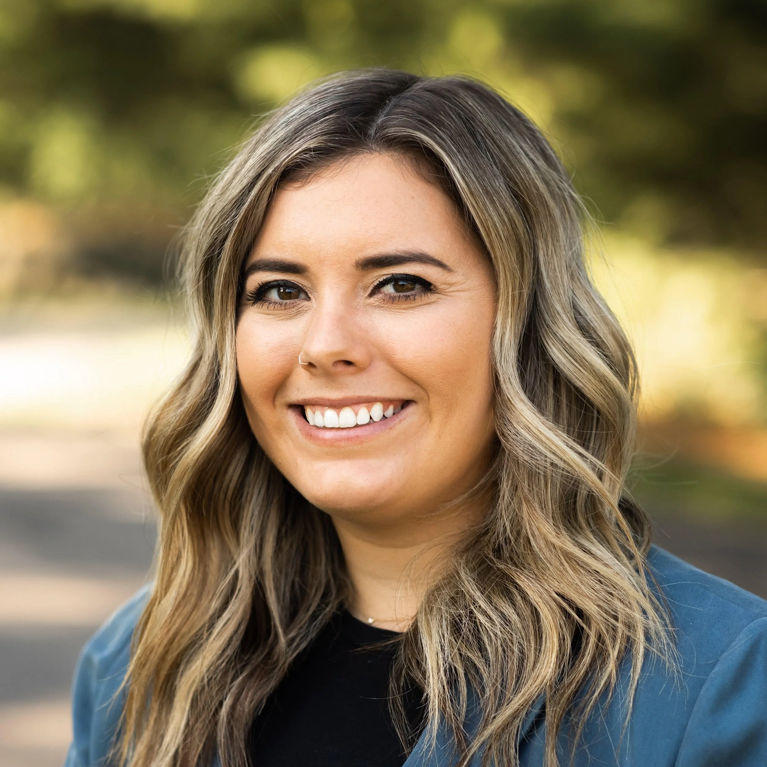 Close-up of a smiling woman with wavy blonde hair and a nose piercing outdoors.
