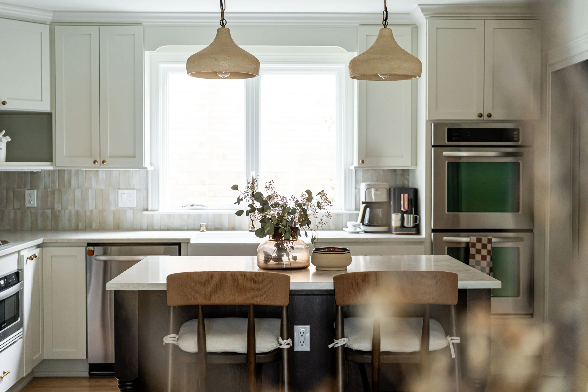 Modern kitchen with white cabinets, stainless steel appliances, a large window, a kitchen island with a flower vase, and hanging pendant lights.