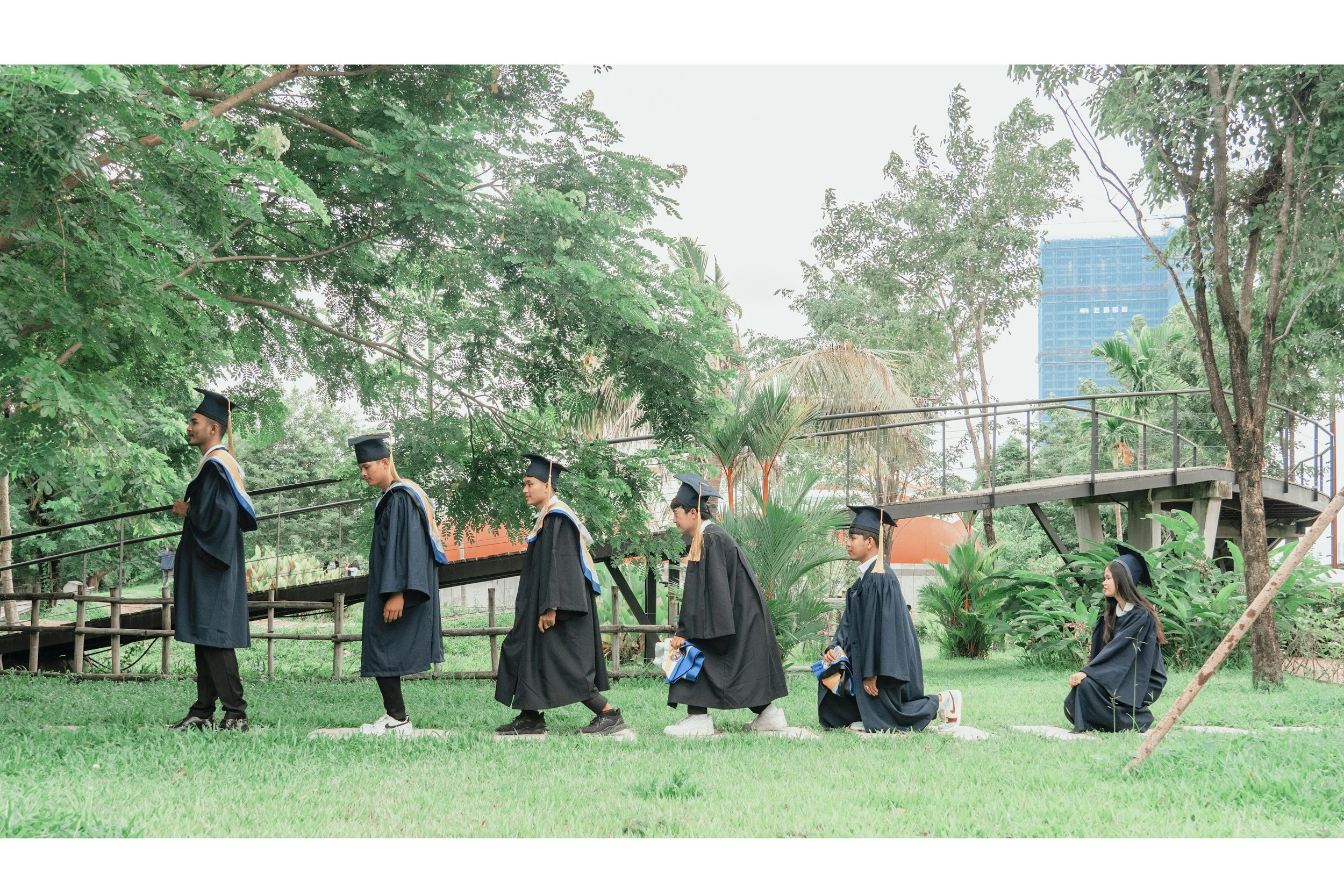 Group of graduates in caps and gowns walking outdoors in a park with lush green trees and a city skyline in the background.