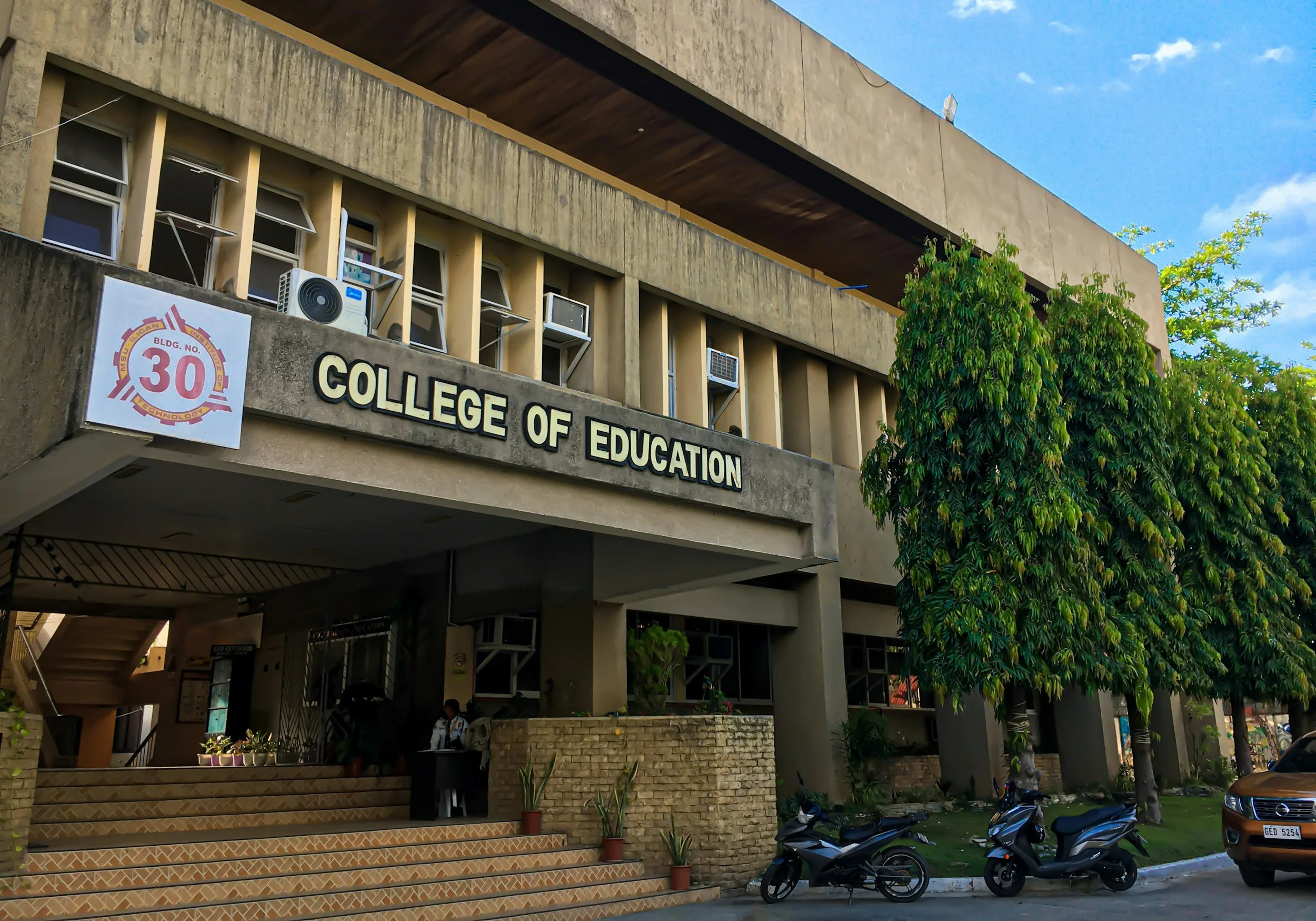 Exterior of a building with a sign reading 'College of Education', some potted plants, motorcycles parked in front, and a tree with green foliage. The building has a concrete facade, several air conditioning units, and a staircase visible underneath.