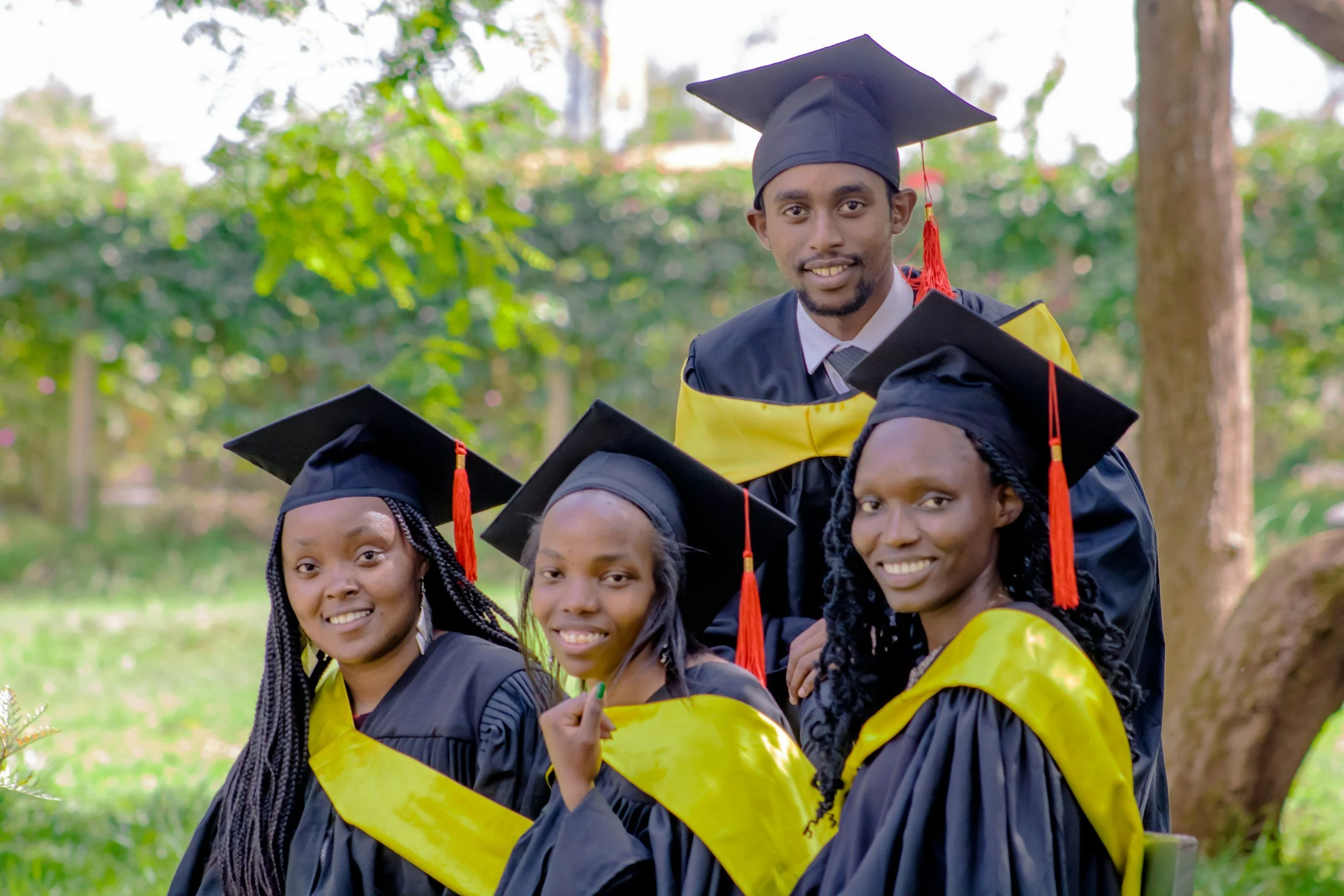 A group of students wearing graduation caps and gowns, smiling outdoors with a background of green trees and sunlight.