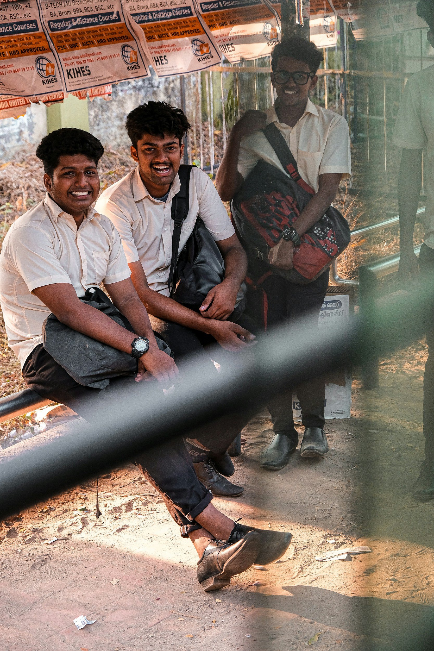Three young men in cream shirts with backpacks, sitting and standing at a bus stop, smiling. Orange advertisement banners above them, dirt ground with some trash, trees visible in the background.