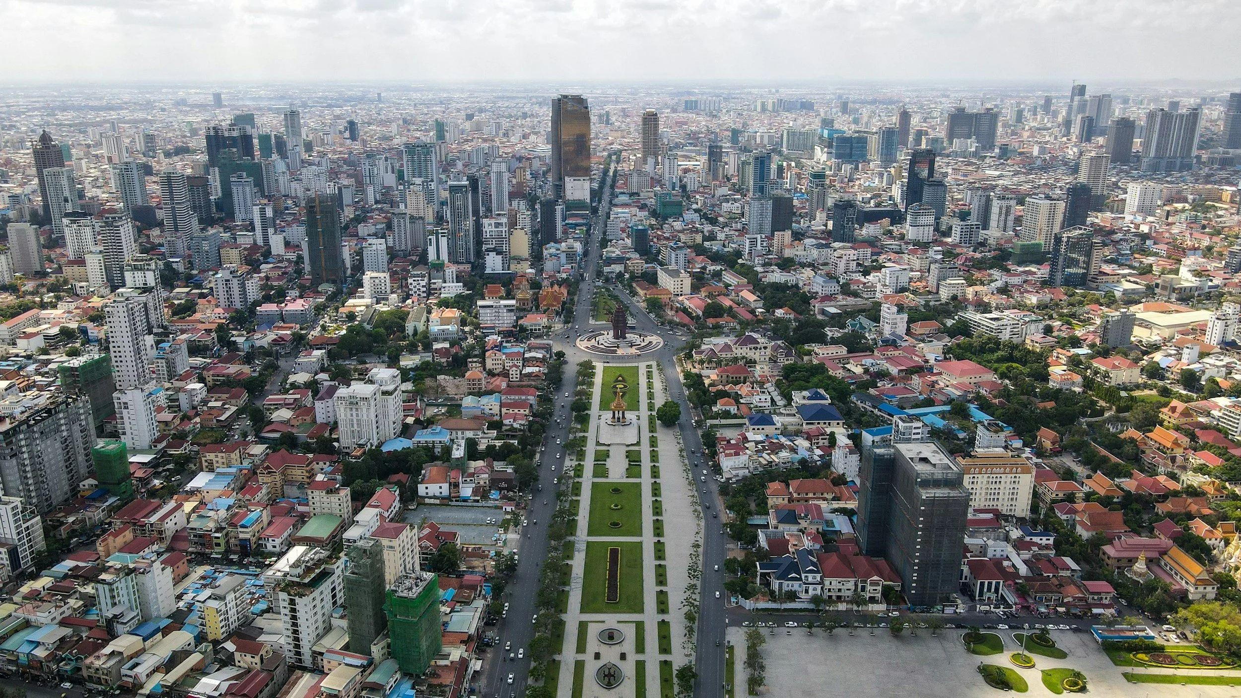 Aerial view of a large city with many high-rise buildings, a central park with a statue and flowerbeds, and densely packed low-rise residential and commercial buildings extending to the horizon.