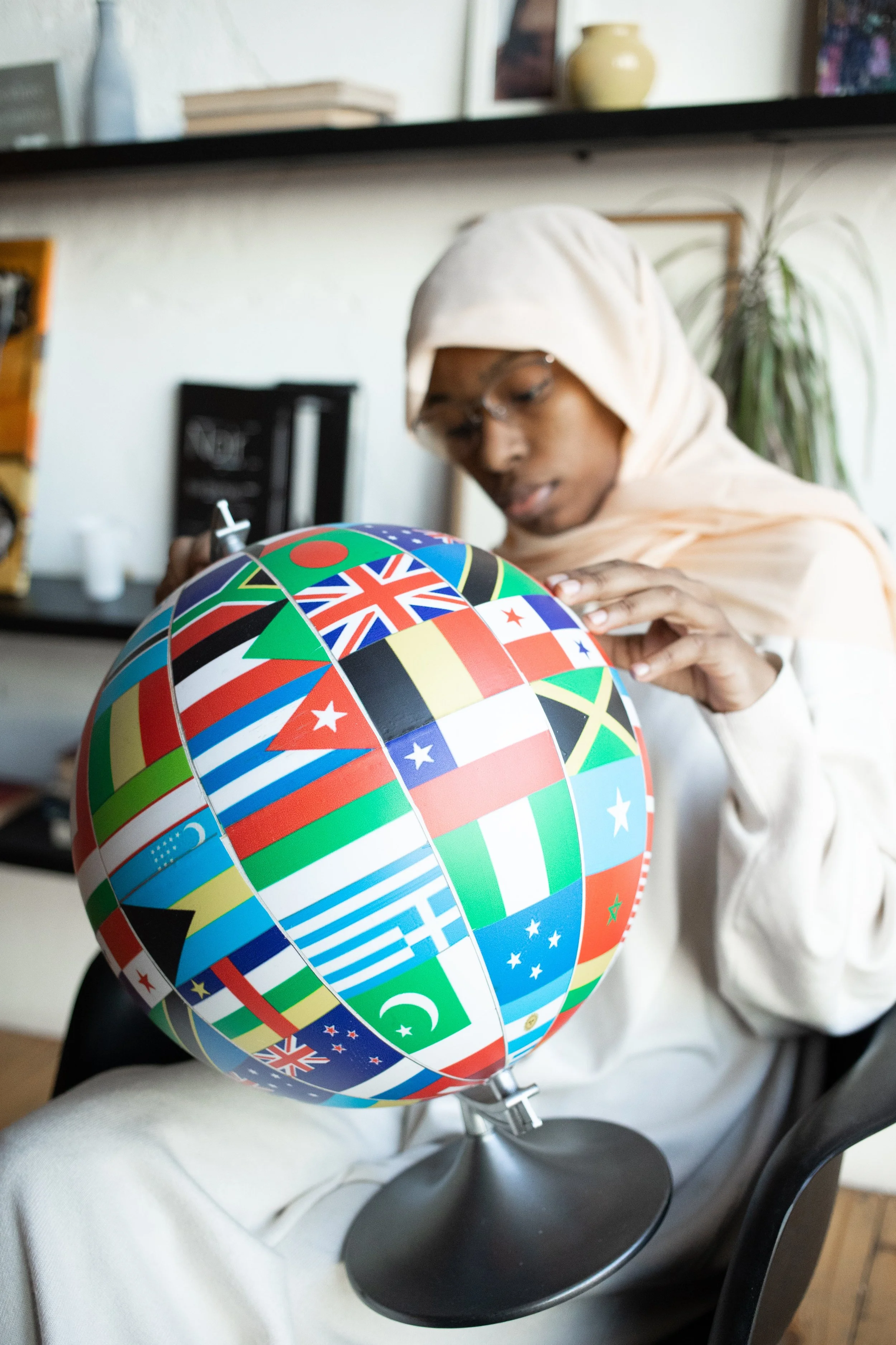 A person wearing a light-colored hoodie and glasses is sitting on a chair, focused on a colorful globe with flag designs. The globe is resting on a table with a white cloth and has a black stand.