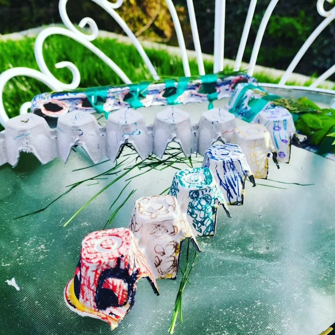 Colorful caterpillar decorated egg carton's on a metallic table outdoors , with a white ornate garden chair in the background and greenery.