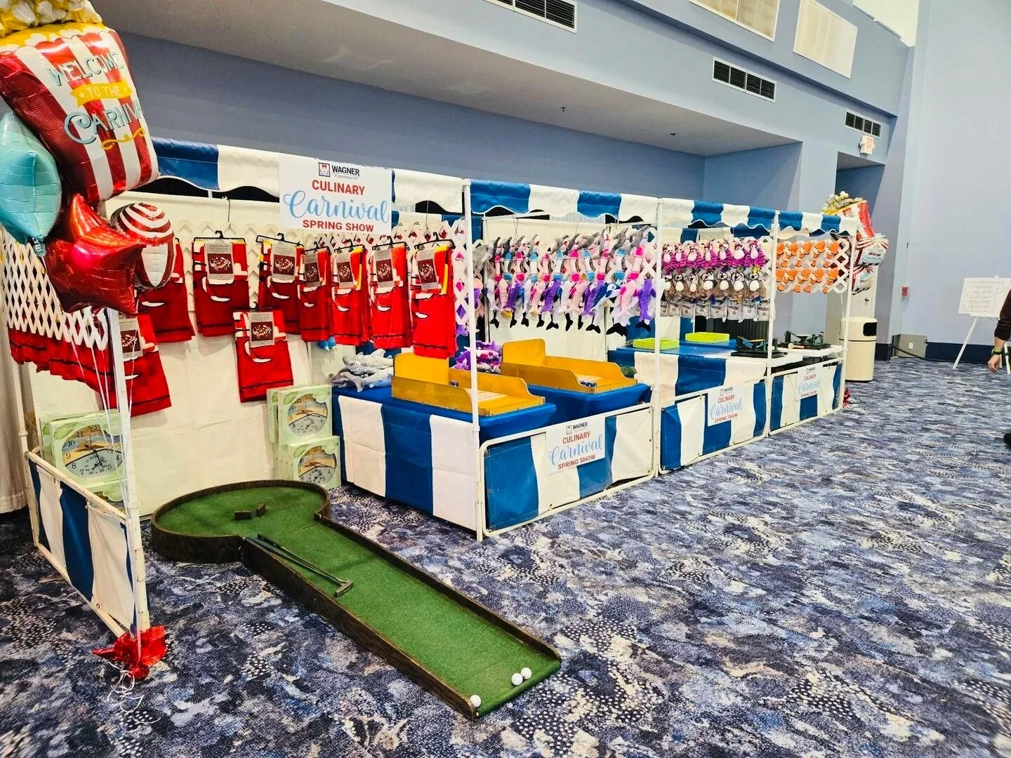 Carnival game booth with plush toys, red aprons, and a mini golf putting green at the Wagner Culinary Carnival Spring Show.